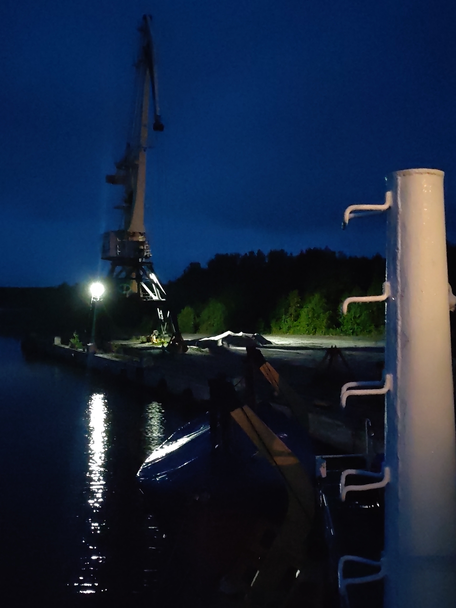 The image, taken at night, shows a port scene in Shalsky, Russia. A large white crane dominates the background, illuminated by a street lamp at its base. A body of water, reflecting light, occupies the left side of the frame. Dock structures extend from the shore, and trees line the horizon. In the foreground, a white cylindrical structure with ladder-like protrusions is visible. A partially obscured blue and yellow boat is also present, suggesting its location near the dock. The sky is a dark blue, indicating nighttime.