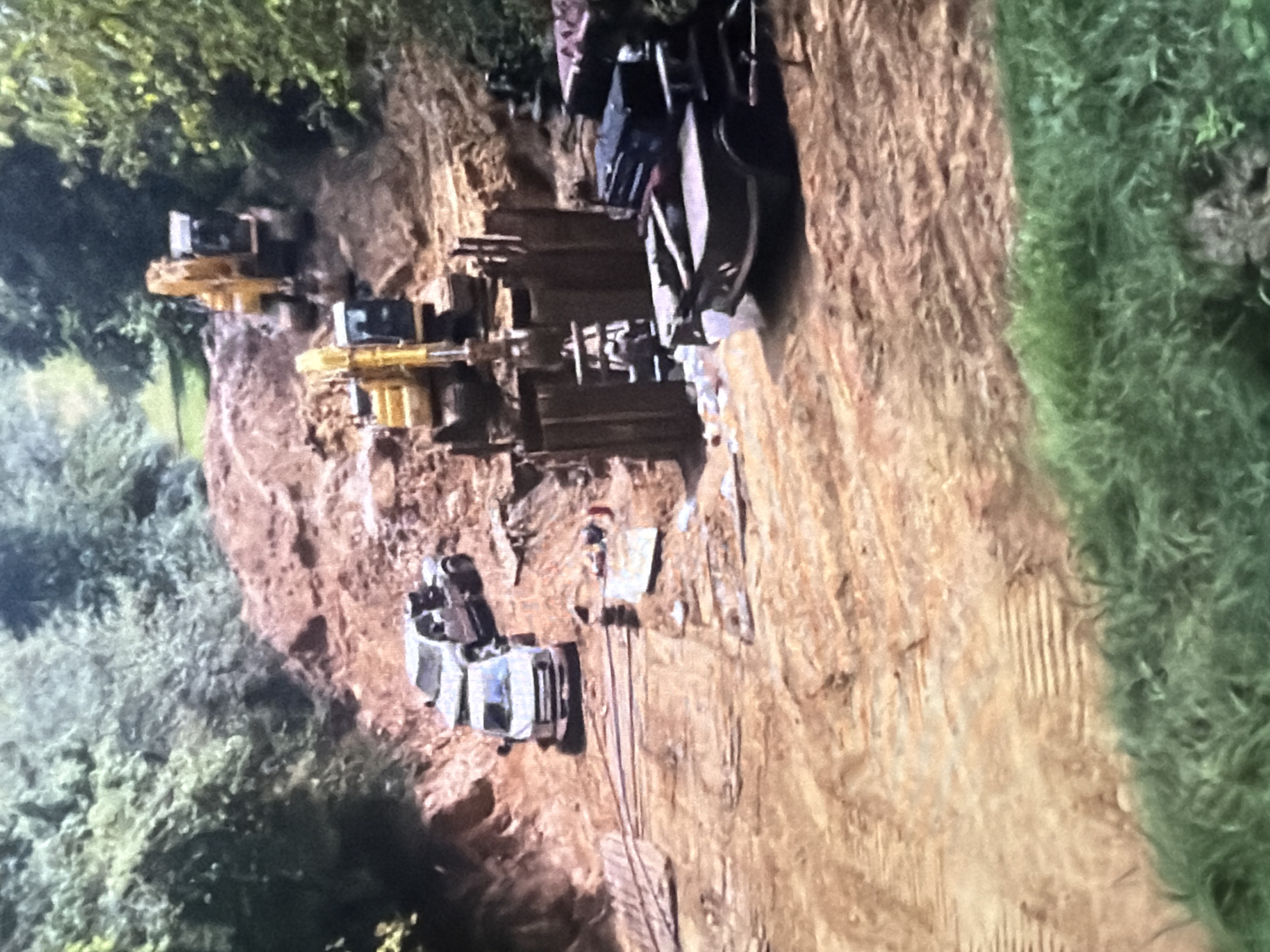 The image depicts a construction site in a wooded area. In the foreground, a flatbed truck with a dark toolbox on its bed is parked on a dirt path. To the left, a white van and a pickup truck are parked on a dirt incline. Further up the incline, two yellow excavators are visible, one actively digging in a pile of dirt. Wooden planks and other construction materials are scattered around the site. The environment is characterized by dirt, mud, trees, and lush green grass. The overall impression is of ongoing earthmoving and construction work. The specific location within the city of Unknown, Unknown, is not discernible from the image.