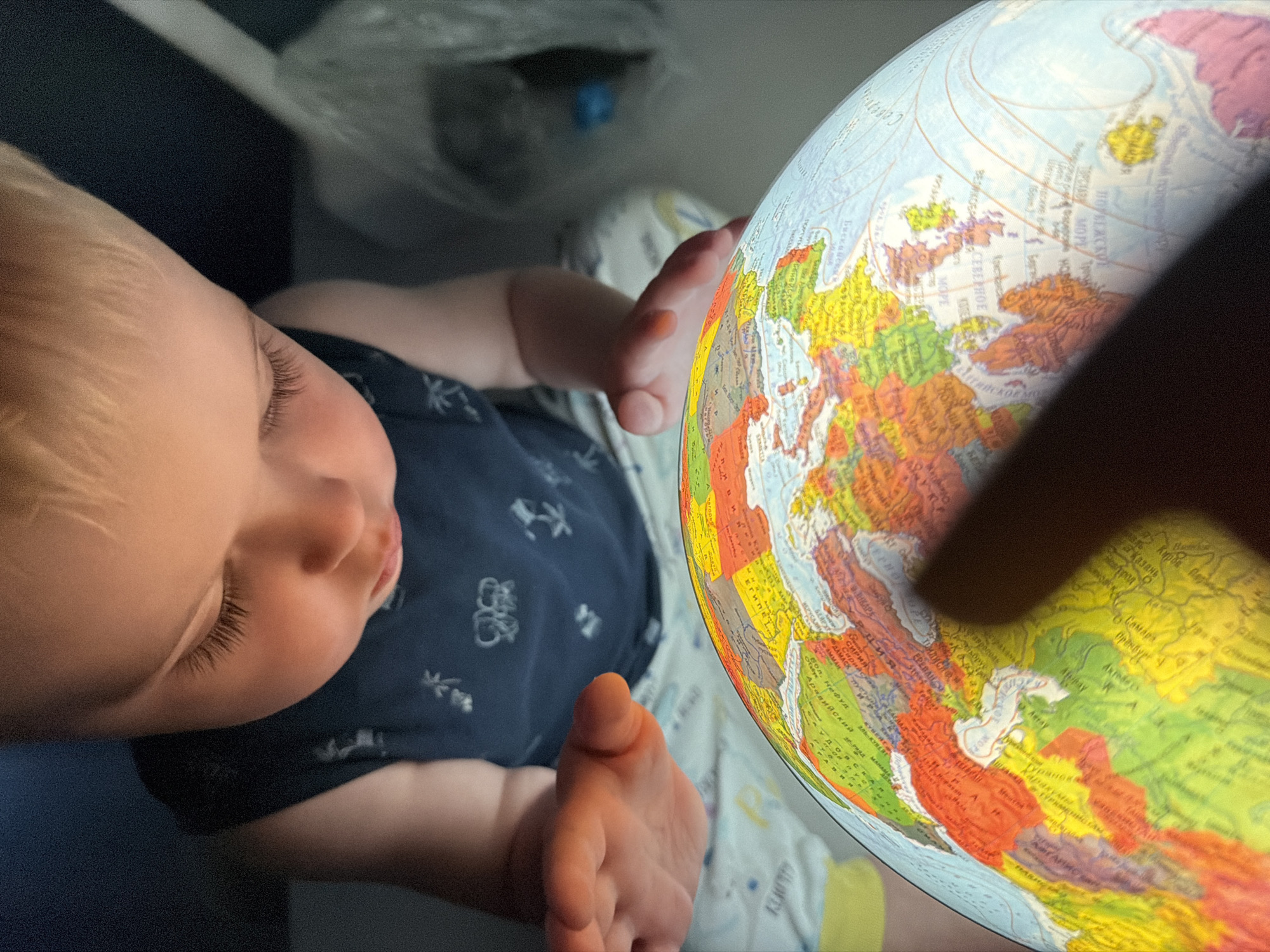 The image shows a close-up, overhead view of a baby's head and arm as they reach towards a brightly lit globe. The baby, with fair hair and closed eyes, appears to be very young, possibly a toddler, and is wearing a dark blue shirt with a subtle pattern. The baby's left hand is extended, with fingers slightly curled, as if about to touch the globe. The globe itself is illuminated from within and displays a detailed map of Europe and parts of Asia. Countries are color-coded, and place names are visible in Cyrillic script. The background is dark, suggesting indoor lighting, and hints of a blanket or fabric are visible. A finger, possibly belonging to an adult, casts a shadow over a portion of the globe, adding a sense of scale and interaction. The text visible on the globe includes country names like "Сирия" (Syria), "Египет" (Egypt), and regions like "Каракумы" (Karakum Desert). The overall impression is one of a gentle moment of exploration and learning, with the baby interacting with a world map. The context of Votkinsk, Russia, is not directly evident from the visual information in the image but is provided as additional information.