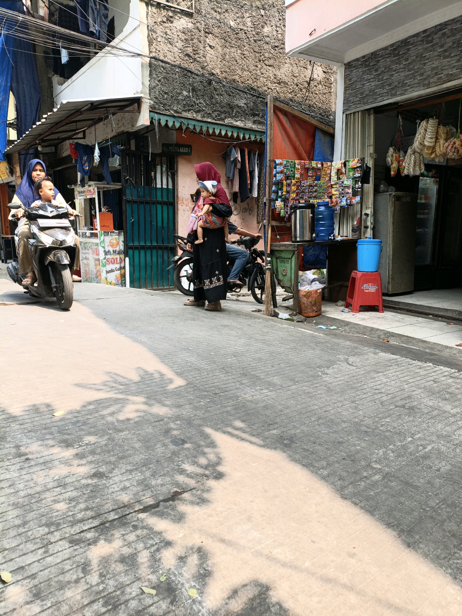 This image captures a street scene in West Jakarta, Indonesia, characterized by a narrow road paved with concrete, marked with visible lines. Sunlight casts dappled shadows across the pavement, suggesting a bright day.
On the left side of the road, a woman wearing a blue hijab and a light-colored shirt is riding a black scooter, with a young child in front of her, also wearing a blue garment. The scooter appears to be in motion, moving away from the viewer. Behind them, a building with a white facade and a corrugated metal roof is visible, with laundry hanging from lines.
To the right of the scooter is a stall or small shopfront. A woman wearing a red hijab and a black garment is standing, carrying a baby on her back. She is positioned near another scooter. The stall displays various items for sale, including bags of snacks and beverages. A large blue water jug and a silver dispenser are prominent items on the stall. In front of the stall, a red stool with the word "Sedap" printed on it is placed, topped with a blue bucket. A trash can with some litter is also present near the stall.
The buildings in the background appear to be densely packed, typical of urban residential areas. One building has a textured stone facade, while another has a more modern, light-colored upper level. The overall impression is of a bustling, everyday street life in a residential and commercial district.