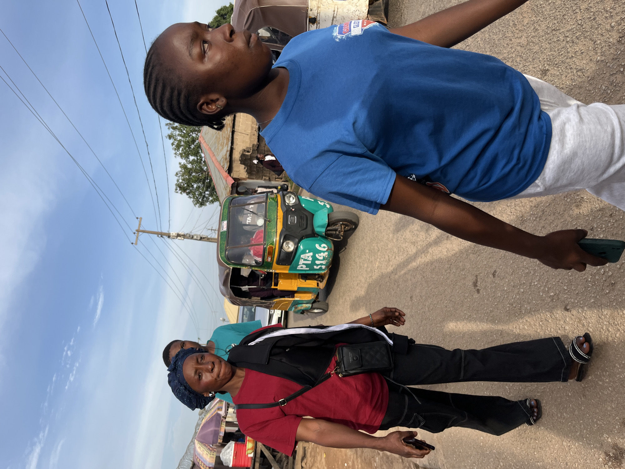 This daytime image captures a vibrant street scene, likely in Jos, Nigeria, featuring two prominent women and a tricycle taxi. The photograph is oriented vertically, creating an unusual perspective.
In the foreground, an older woman stands or walks, looking towards the viewer with a slight, friendly smile. She is dressed in a red short-sleeved t-shirt, black pants, and a black jacket or vest worn over the shirt. A black crossbody bag with a quilted pattern hangs over her shoulder, and she holds a dark-colored mobile phone in her right hand. Her head is covered with a dark blue patterned headwrap.
To her right and slightly behind, a younger woman is depicted in profile, looking upwards and to her left (from the viewer's perspective). She wears a bright blue t-shirt, featuring two patches on the shoulder area; one clearly reads "GLEX" and another appears to be "GLO". She is also wearing white sweatpants. Her hair is styled in neat braids, and she has a small hoop earring. Her left arm is extended downwards, holding a dark-colored smartphone.
In the mid-ground, a green and yellow tricycle taxi, commonly known as a "kuk-kure," is visible. The identification number "PTA-5146" is prominently displayed on its front. The tricycle appears to be either parked or moving slowly on the paved road.
The background reveals a bustling environment with additional, indistinct figures, market stalls or canopies, and buildings with corrugated metal roofs. A network of utility poles and overhead power lines crisscrosses the clear blue sky, which shows a few scattered clouds. The overall impression is one of daily life in a lively community under bright, sunny conditions.