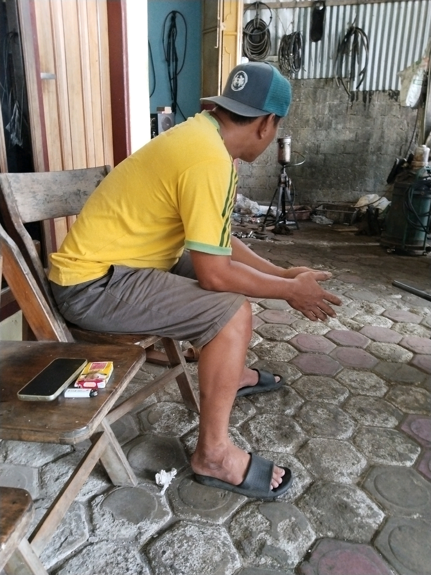 The image captures a man wearing a yellow t-shirt and gray shorts, seated on a wooden chair. He has a dark baseball cap on his head and is wearing dark gray slide sandals. In his lap, he holds a pack of cigarettes and a lighter. On a small wooden table beside him, a smartphone is placed. The man's hands are resting in front of him on the patterned concrete floor, which appears to be an outdoor or semi-outdoor area. In the background, a corrugated metal wall is visible, with various objects hanging on it, suggesting a workshop or storage area. The overall setting resembles a casual, perhaps industrial, environment. The location is identified as Prigi, Indonesia.