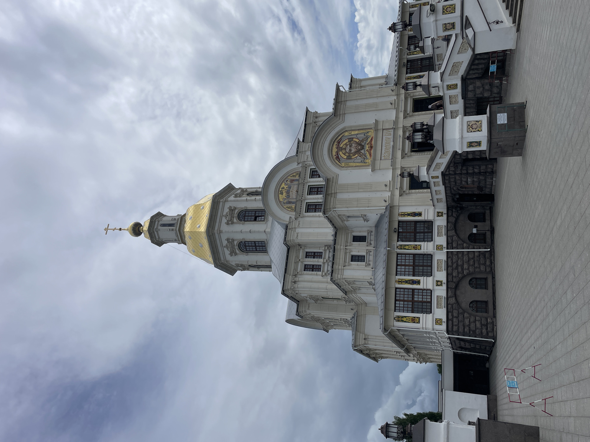 The image shows the Trinity Cathedral in Diveyevo, Russia, a large, white, ornate building with a golden dome topped with a cross. The cathedral is under a cloudy sky. The foreground is a paved courtyard with a red and white barrier. The building has multiple windows and decorative elements, including mosaics on the facade. Some people are visible near the entrance. The architecture suggests a religious or historical site.