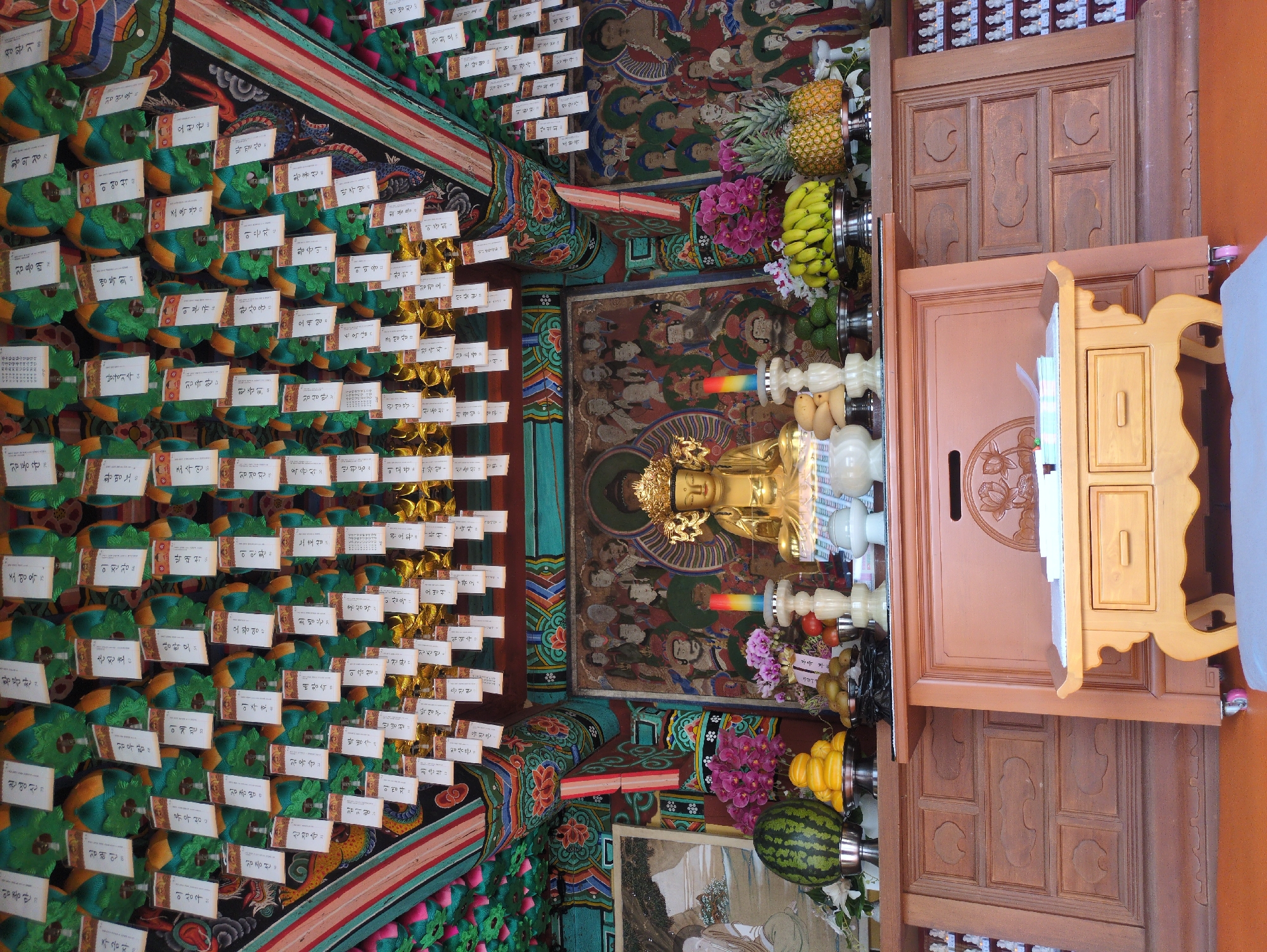 This is an indoor shot of a Buddhist temple in Gongju-si, South Korea. The image is rotated counter-clockwise so the viewer sees a horizontal orientation. The ceiling is adorned with ornate paintings and rows of small green and gold ornaments with white labels written in Korean. A golden Buddha statue sits on a table with offerings of fruits, including pineapples, bananas, and a watermelon. A wooden cabinet and a smaller yellow table are next to the Buddha. The setting is richly decorated, reflecting the spiritual nature of the location.