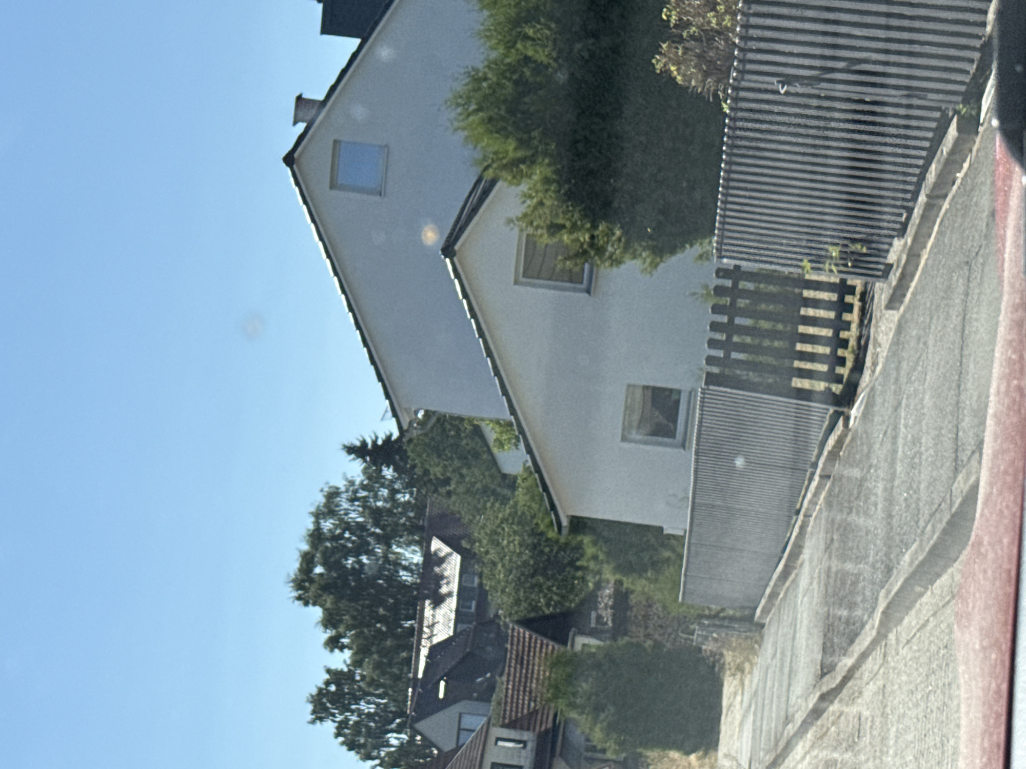 The image shows a street scene in Bad Sachsa, Germany, under a clear blue sky. Two houses are visible, one larger and white with a gable roof and a small square window, and another smaller white house with a window. Both houses are surrounded by greenery, including trees and bushes. A grey metal fence with vertical slats runs along the street. A paved pathway or driveway slopes upwards from the foreground. The overall impression is a residential area on a sunny day.