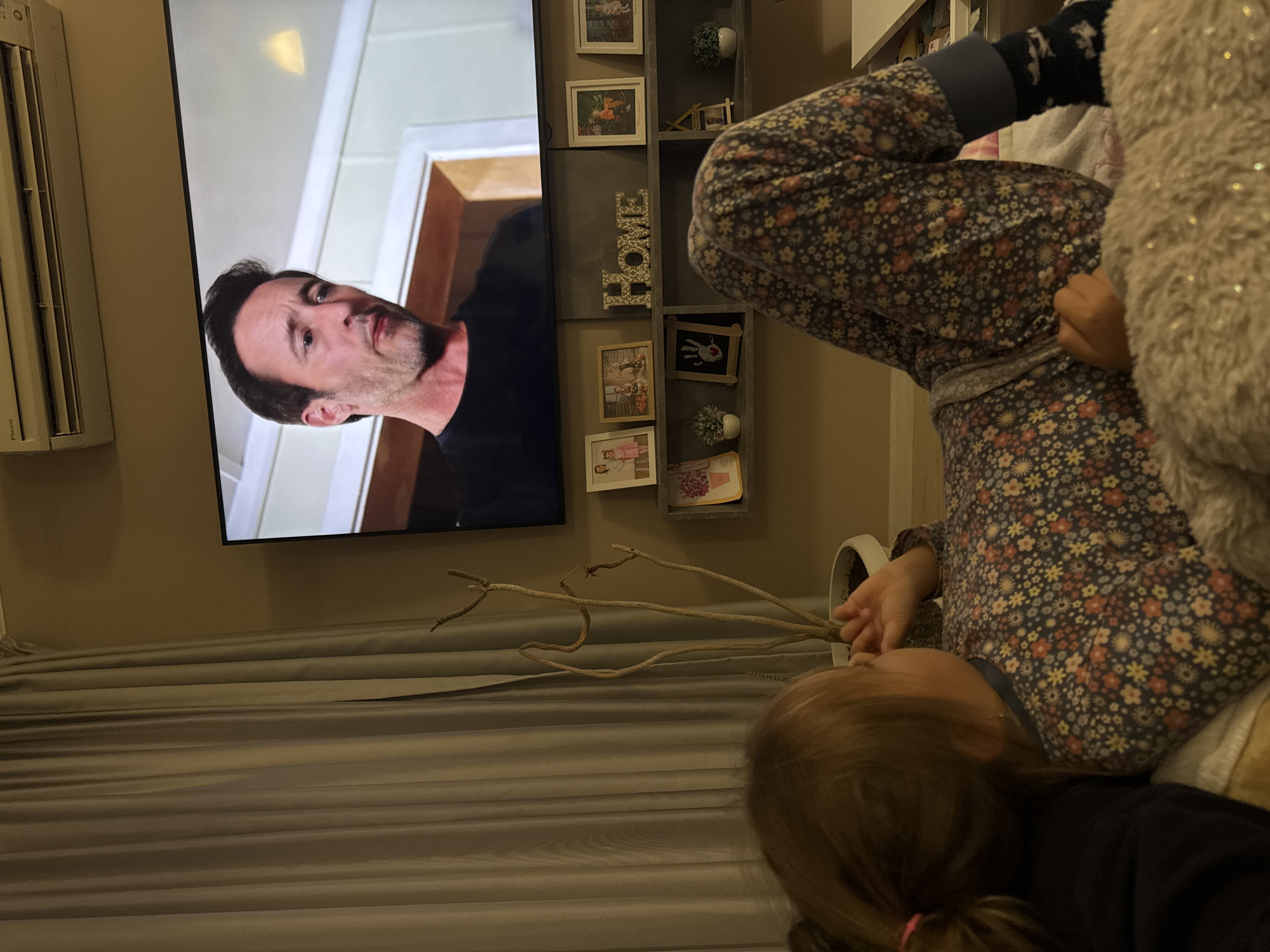 A child is lying in an upside-down position in a domestic setting, partially obscured by a fluffy material, possibly a blanket. The child is wearing floral print clothing. A television is mounted on the wall, displaying a man. There is a shelf with framed pictures and a decorative "HOME" sign next to the television. The background includes grey curtains and decorative branches in a white vase. There is no information to determine a location in Varna, Bulgaria.