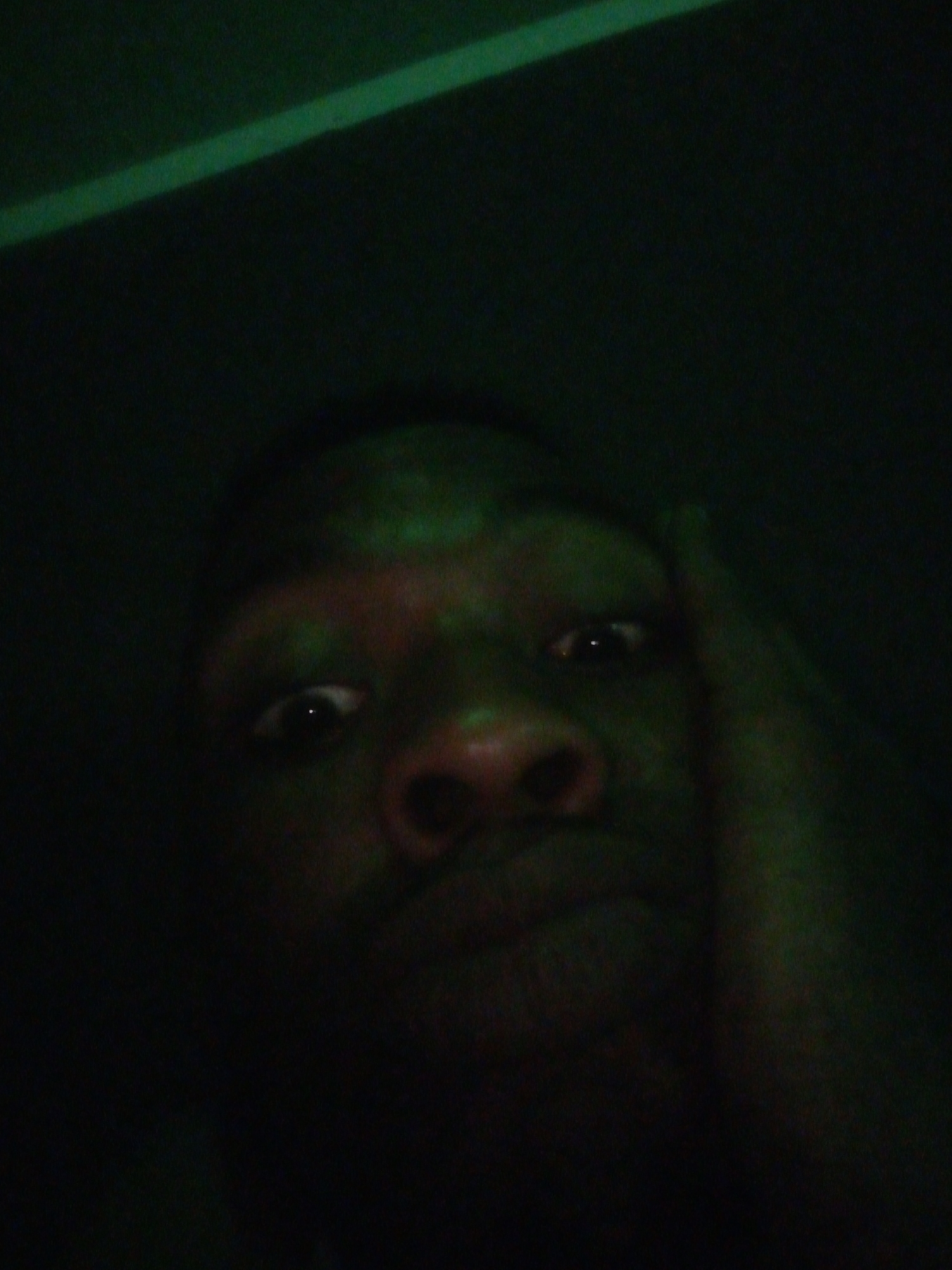 A dimly lit selfie captures a person with dark skin. The person's right hand is positioned near their face. A faint green glow is present on the subject's skin, as well as a strip of green light in the background. The environment appears to be dark and undefined.