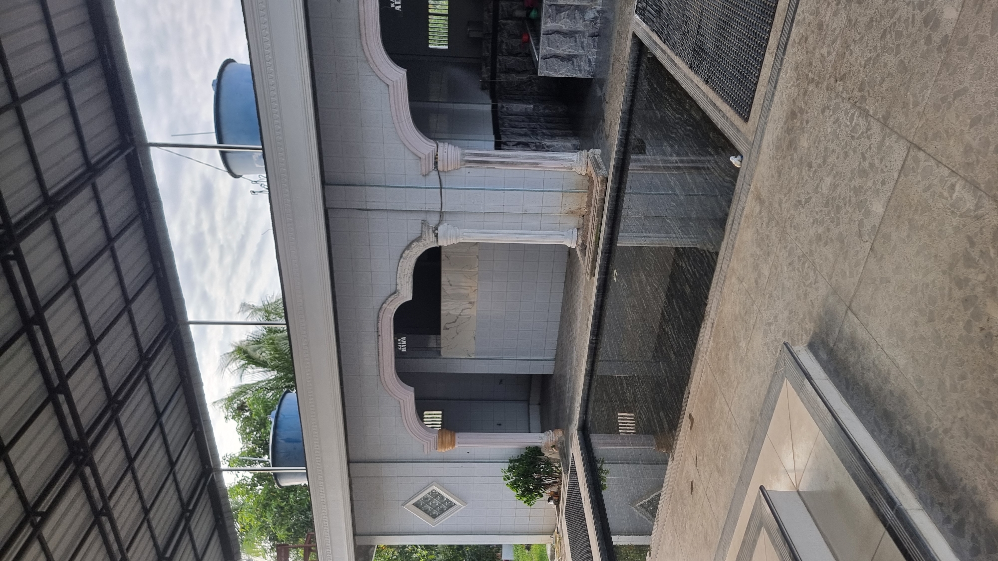The image shows an outdoor ablution area with a shallow pool of dark water in the foreground. The pool is rectangular and surrounded by tiled paving. To the left of the pool are a set of tiled stairs. Behind the pool is a low-rise building with white tiled walls and decorative arches. Two large blue water tanks are visible on the roof of the building. The scene appears to be located in Meunasahlhok, Indonesia, given the context provided. The sky is overcast. There are no people visible in the image.
