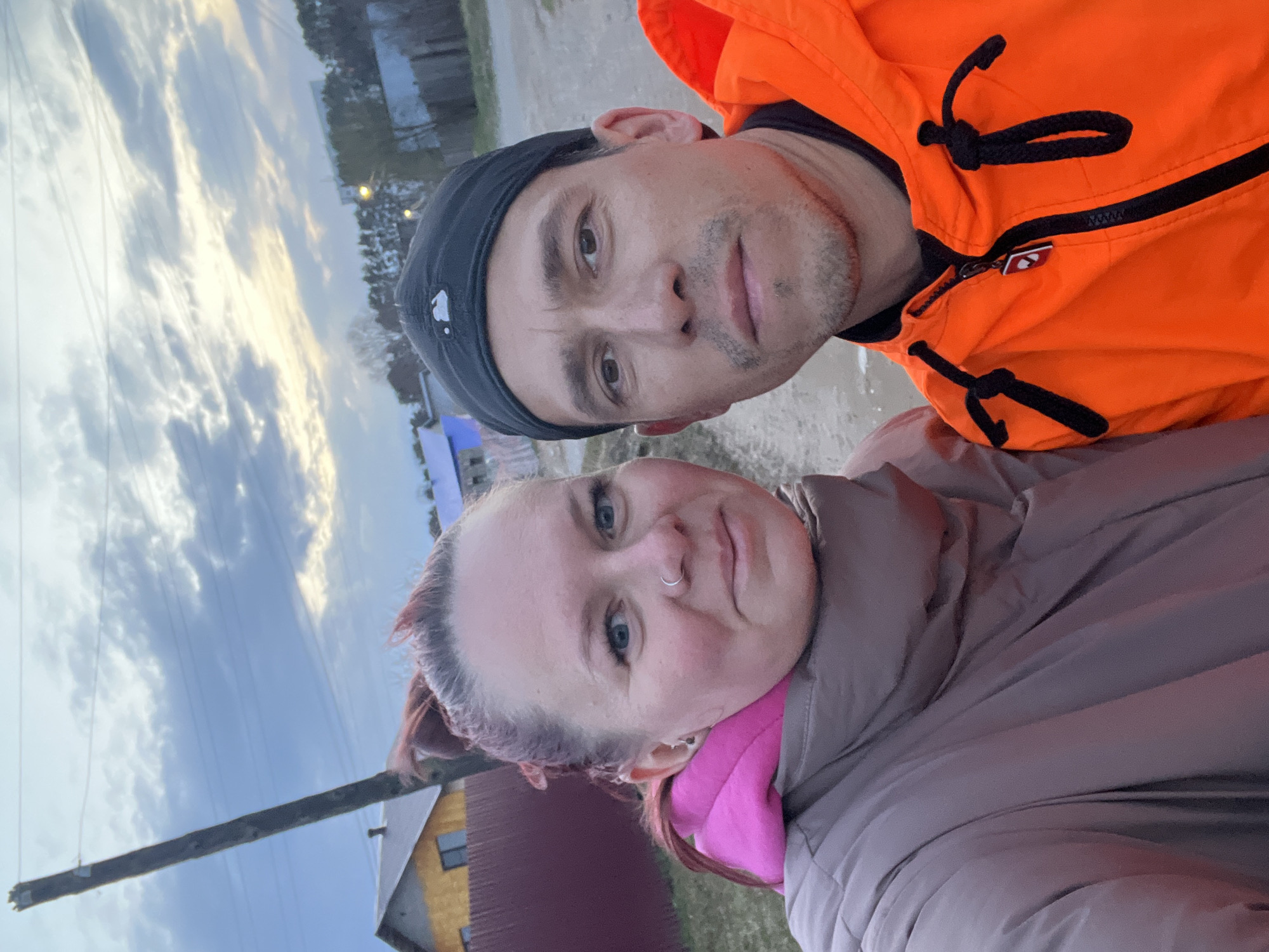 The image is a selfie of a man and a woman in Chaikovsky, Russia. The man is wearing a black cap and an orange jacket. The woman has reddish hair and is wearing a tan jacket with a pink lining. In the background are buildings and a cloudy sky. There is a utility pole and wires.