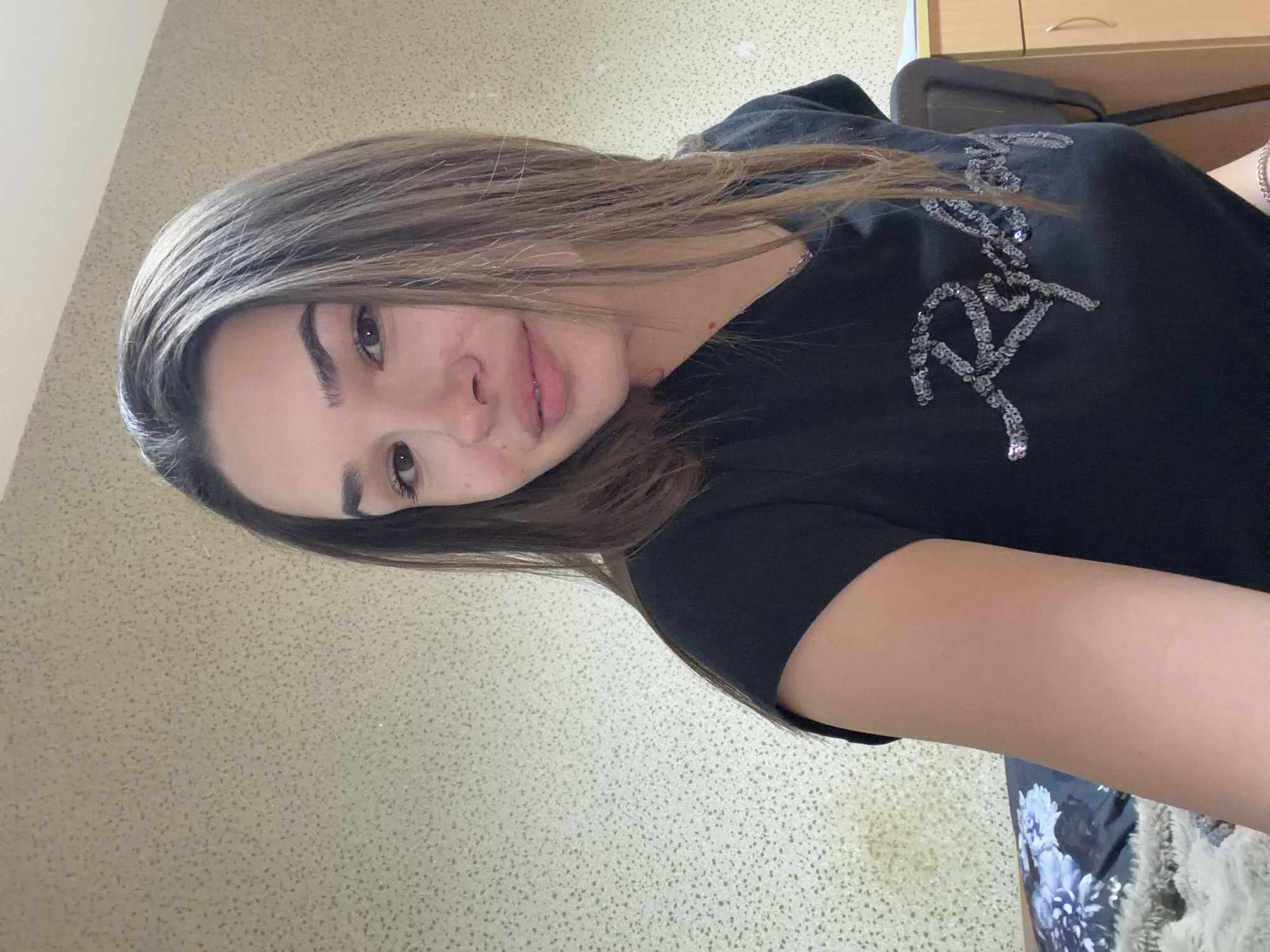 A young woman with long, light brown hair is depicted in this medium shot. Her hair falls over the left side of her face and shoulder. She has fair skin and dark eyebrows and eyes. Her lips are slightly parted. She is wearing a black t-shirt with a glittery logo on the upper right chest. The background is a light-colored wall with a subtle pattern. A piece of furniture, potentially a desk, is visible in the upper right corner. A floral-patterned fabric can be seen in the bottom right. The image appears to be taken indoors, possibly in a home or office environment. There is no information to indicate that the image was taken in Sofia, Bulgaria.