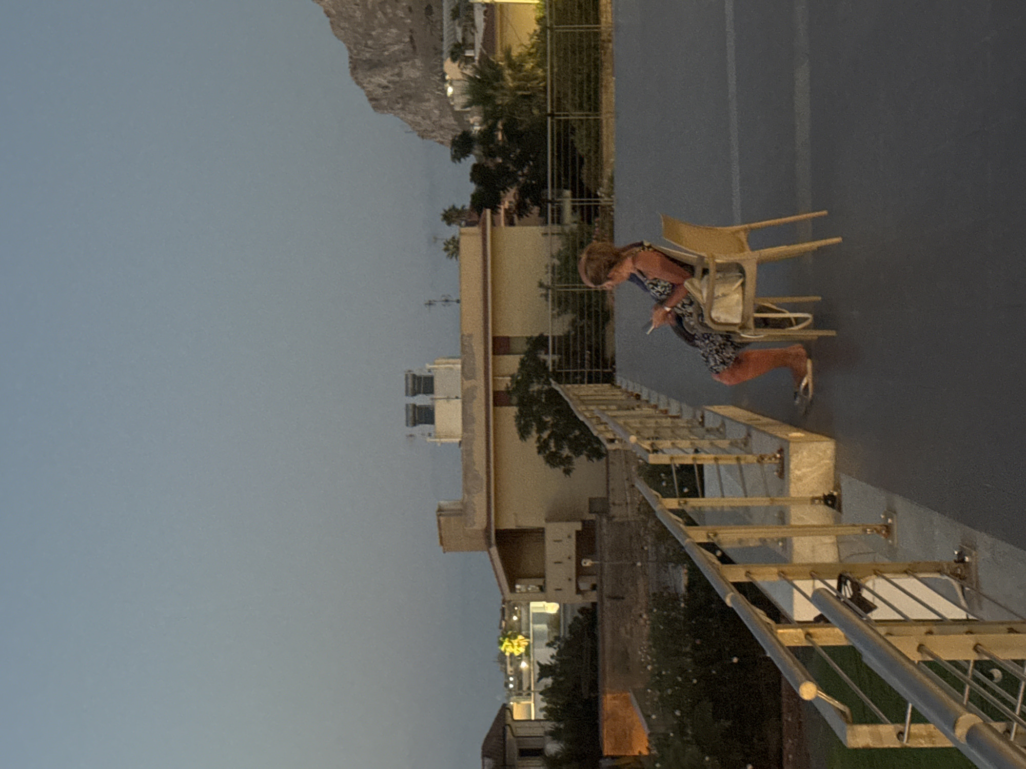A full-shot image captures an outdoor scene at dusk, featuring a woman sitting on a chair on a paved area with a metal railing in the foreground. The woman, dressed in a patterned dress, is holding a mobile phone and appears to be looking at it. She is seated on a light-colored plastic chair. Behind her, a beige building with a balcony and some rooftop structures is visible, along with greenery and trees. Further in the distance, a large rocky mountain dominates the skyline, with lights from buildings scattered below. The sky is a gradient of pale blue and gray, indicating the twilight hour. This scene is characteristic of the coastal town of Isola delle Femmine, Italy, given the architectural style and the presence of the prominent rocky outcrop, which is a distinctive landmark of the area.