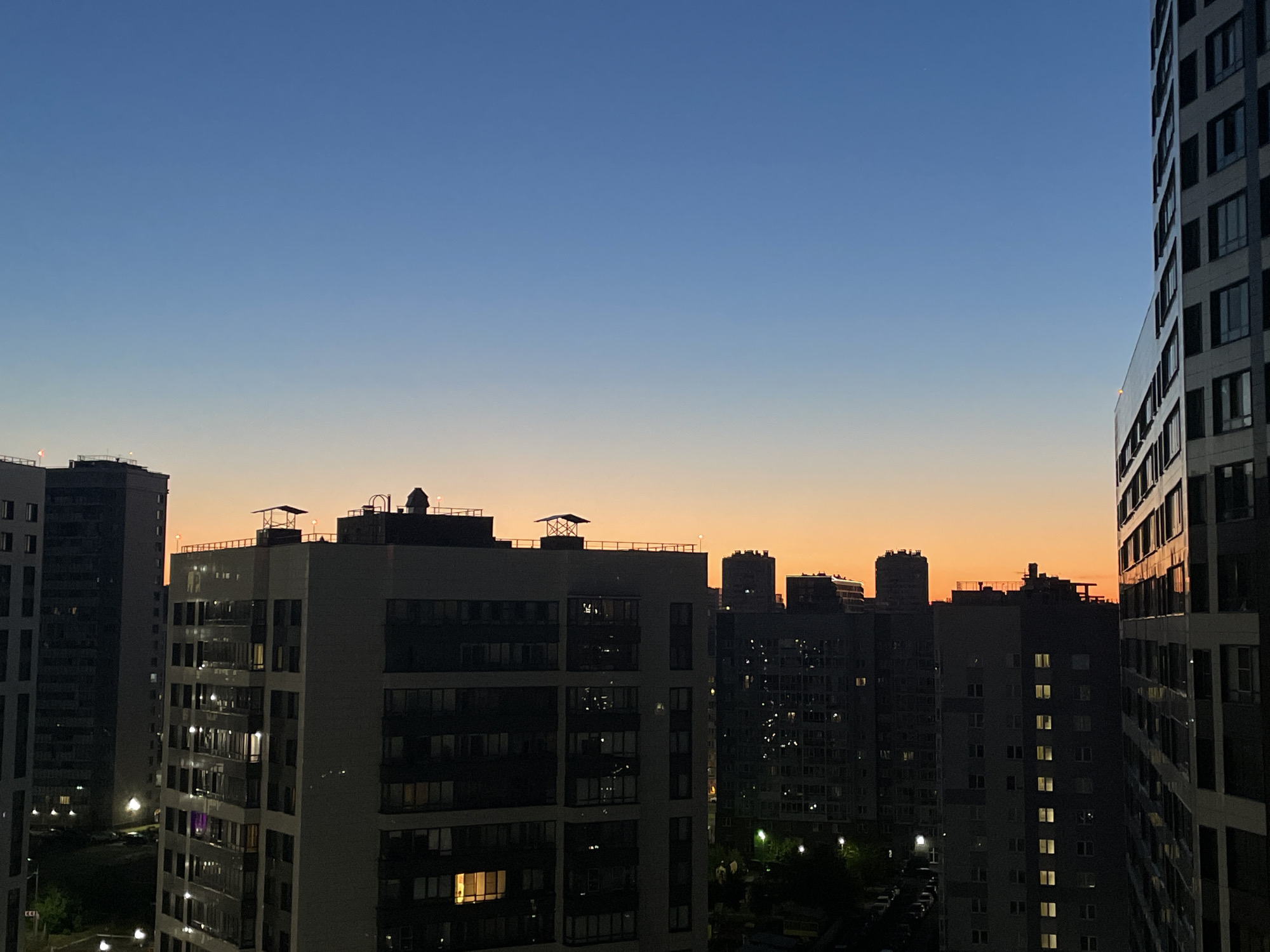 A panoramic, eye-level shot captures a cityscape at dusk in Sinyavino, Russia. The sky transitions from a clear, deep blue at the top to a gradient of soft orange and yellow near the horizon, indicating the setting sun. Dominating the foreground and midground are numerous apartment buildings of varying heights, their facades lit by the warm glow of apartment windows. Some buildings are closer and more detailed, revealing their modern architectural styles with balconies and repetitive window patterns. The building on the far right, a sleek, tall structure, is angled towards the viewer, its windows reflecting the twilight sky. In the distance, the silhouettes of more buildings are visible against the colorful sky. The overall impression is one of urban residential life at the close of day.