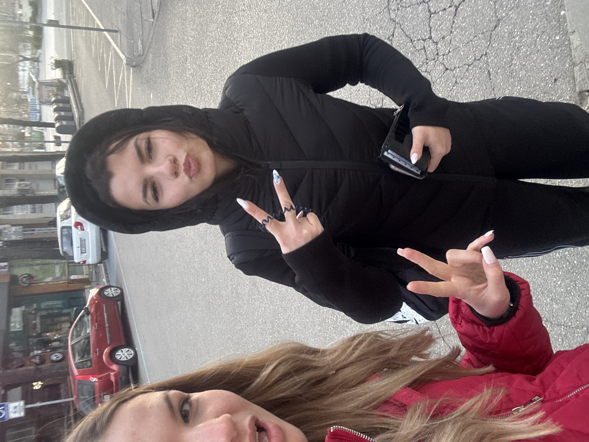 Two young women are posing for a selfie in an urban environment. One woman, wearing a black hooded puffer jacket and black pants, is facing the camera and holding a small black wallet. She is making a peace sign with one hand. The other woman, partially visible at the bottom of the frame, is wearing a red puffer jacket.
In the background, there is a street scene with parked cars, including a red car and a white car. Buildings are visible behind the cars. Based on the street signs and architectural style, the location appears to be in Burgas, Bulgaria. The scene is well-lit, suggesting it is daytime. The surface underfoot appears to be asphalt.
