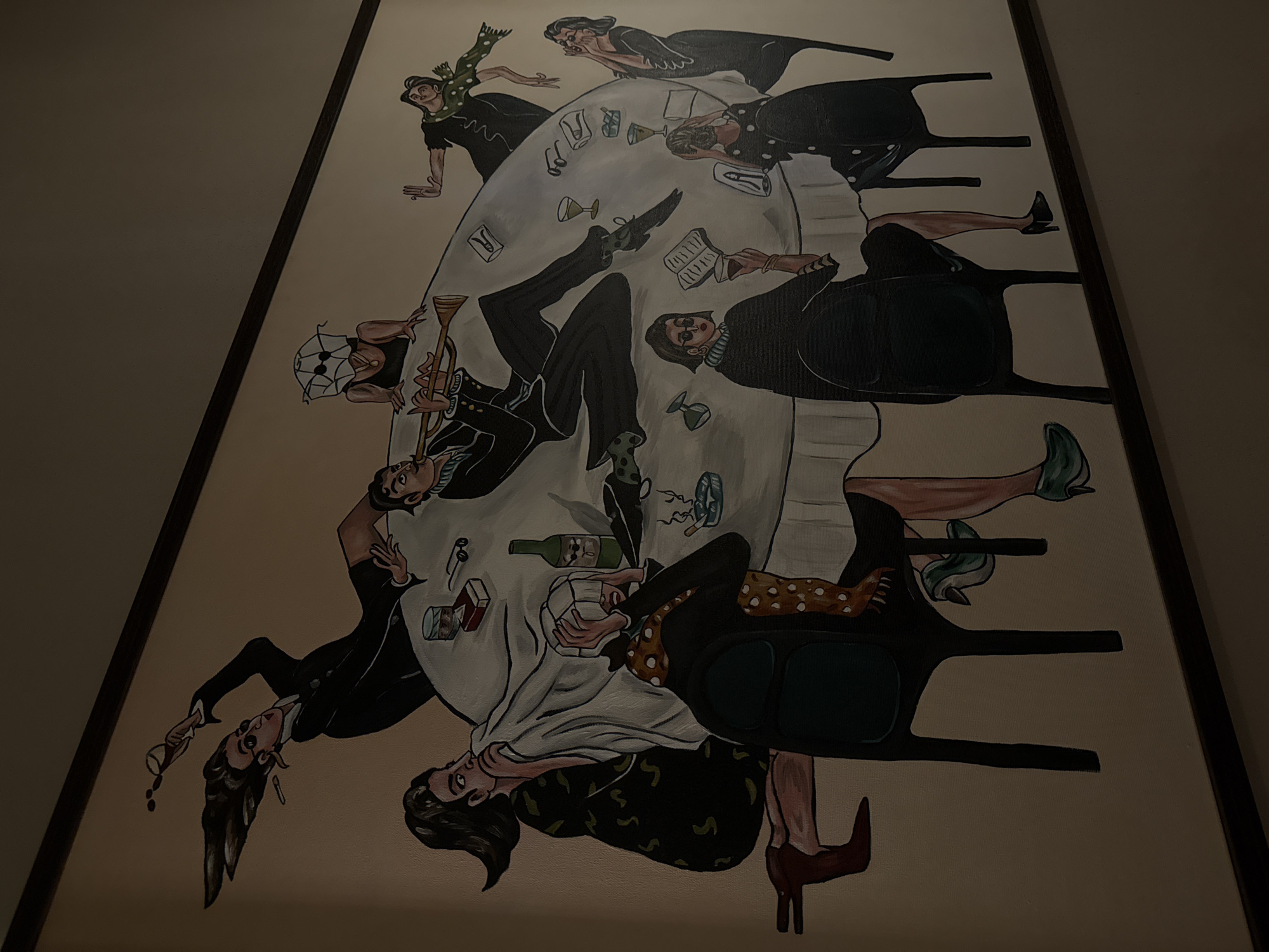 The image is a framed painting on a wall, likely located indoors. The painting depicts a chaotic dinner scene. Around a large, round table covered with a white tablecloth are several figures, seemingly mid-action and in a state of disarray. Some are falling off chairs, others are lying on the table, and various items like glasses, books, and a bottle of wine are scattered across the table. The figures are wearing clothing from different eras, primarily in shades of black and white, with some hints of patterns such as polka dots or floral designs. The style of the painting is somewhat cartoonish and exaggerated, giving the scene a surreal and frantic feel. The lighting is dim, focusing on the painting and casting the surrounding wall in shadow. There is no immediate indication that the painting is specifically located in Yerevan, Armenia.