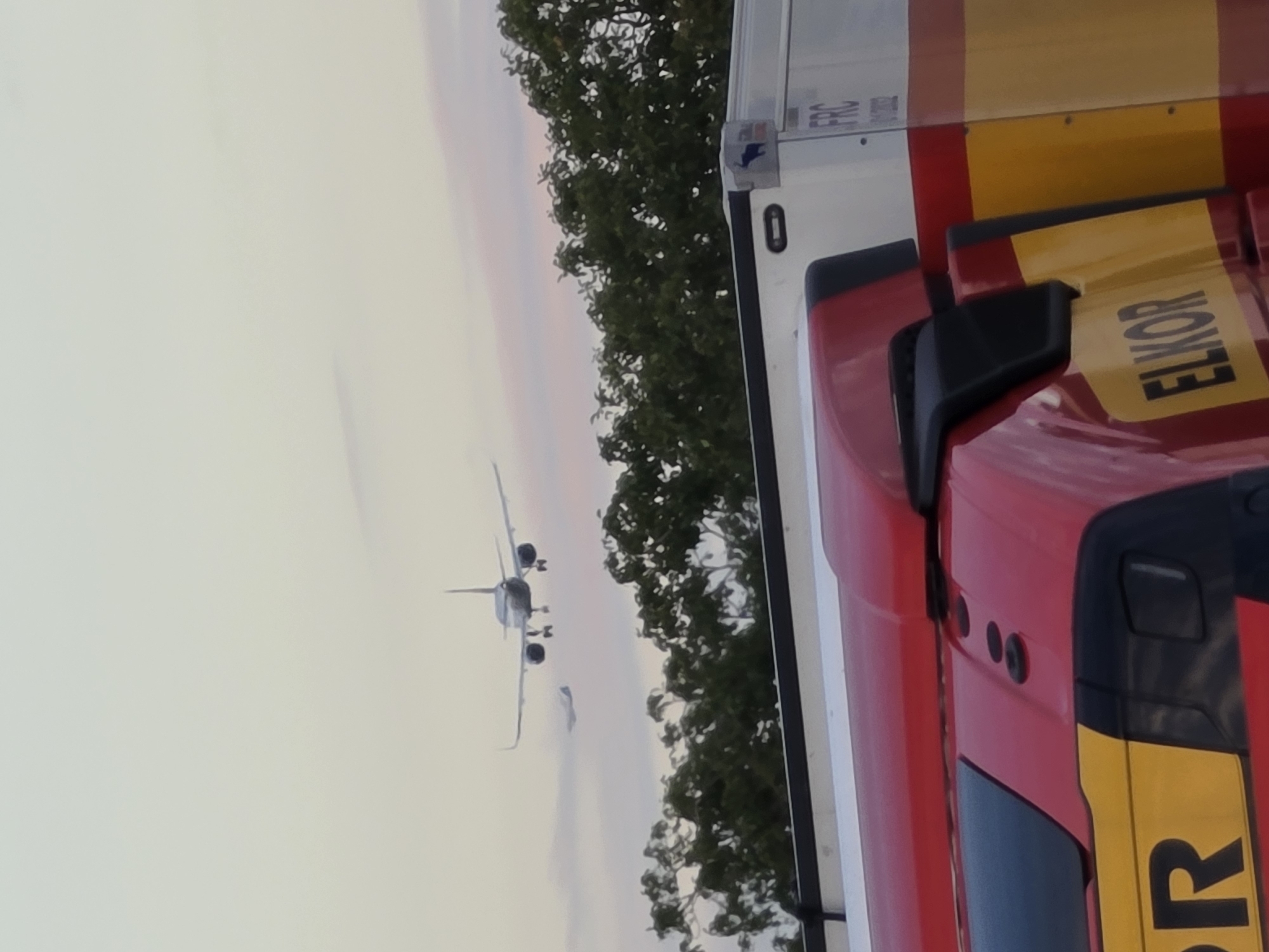 The image shows a red and yellow truck cab in the foreground, with the partial word "ELKOR" visible on a yellow panel. Behind the truck, a line of green trees is visible, and beyond the trees, two airplanes are in the sky. The larger airplane is in the upper center of the image, appearing to be on approach for landing, with its landing gear extended. A smaller airplane is visible to the left and slightly below the larger one. The sky is a pale, hazy color, with hints of pink and orange suggesting either sunrise or sunset. The overall scene appears to be an outdoor location, possibly near an airport or in an area with aerial traffic. The context of Flörsheim, Germany, is not visually evident from the image itself.