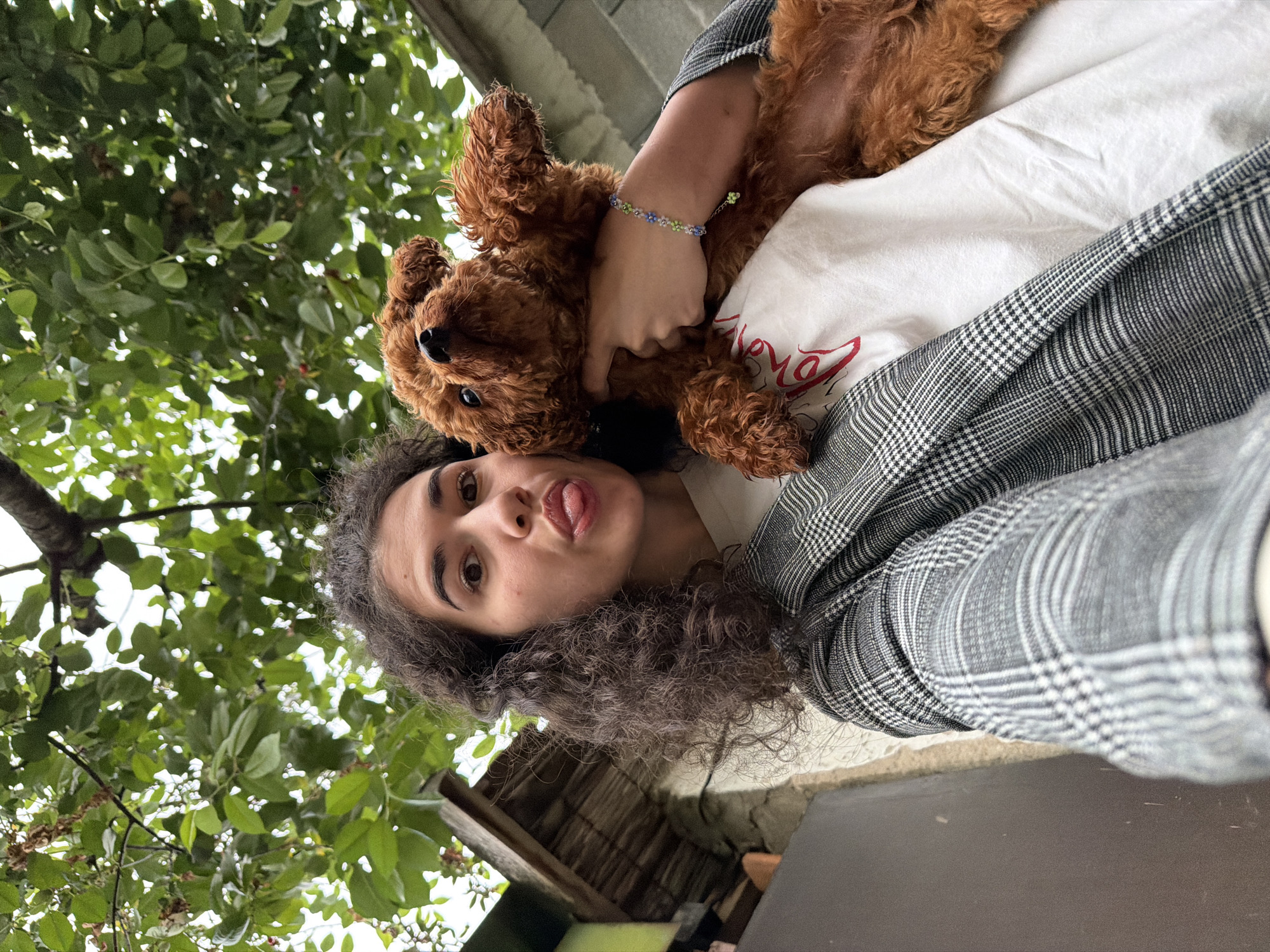 A young woman with curly brown hair, wearing a grey plaid jacket over a white t-shirt, poses for a selfie while holding a small, reddish-brown poodle. The woman is sticking her tongue out slightly. She is wearing a bracelet with blue and green beads on her left wrist. The background shows greenery from trees, and part of a rustic wooden structure and a stone wall, suggesting an outdoor setting in a residential area. The lighting indicates it is daytime. The image quality is clear, allowing for detailed observation of the subjects and their immediate surroundings.