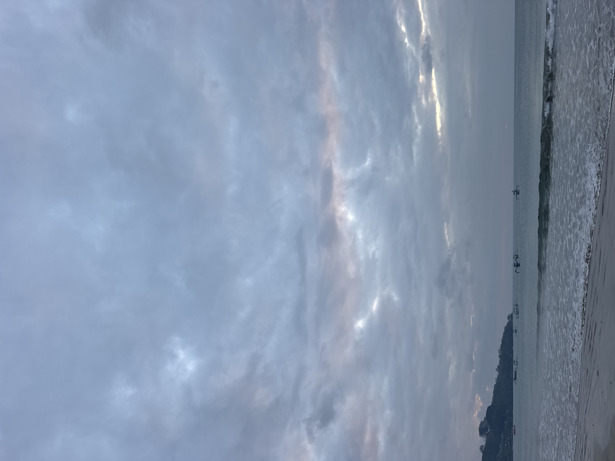 This is a vertical, full-frame, eye-level shot of a beach scene in Kuta Selatan, Indonesia, likely taken at dusk or dawn due to the muted lighting and cloudy sky.
The main subjects are the sky, the ocean, and a small section of beach. The sky is dominated by an expansive layer of overcast clouds, with hints of soft pink and yellow light breaking through in some areas, suggesting either sunrise or sunset. The sea is calm, with gentle waves lapping at the shore. The water is a muted grey-blue, reflecting the overcast sky. A sandy beach occupies the right side of the frame, with the edge of the water creating a foamy line of surf. In the distance, several small boats are visible on the water, and a dark, tree-covered hillside with some structures can be seen along the coastline. The overall mood is serene and tranquil, with a sense of peacefulness evoked by the soft lighting and the gentle sounds of the waves. There is no visible text in the image.