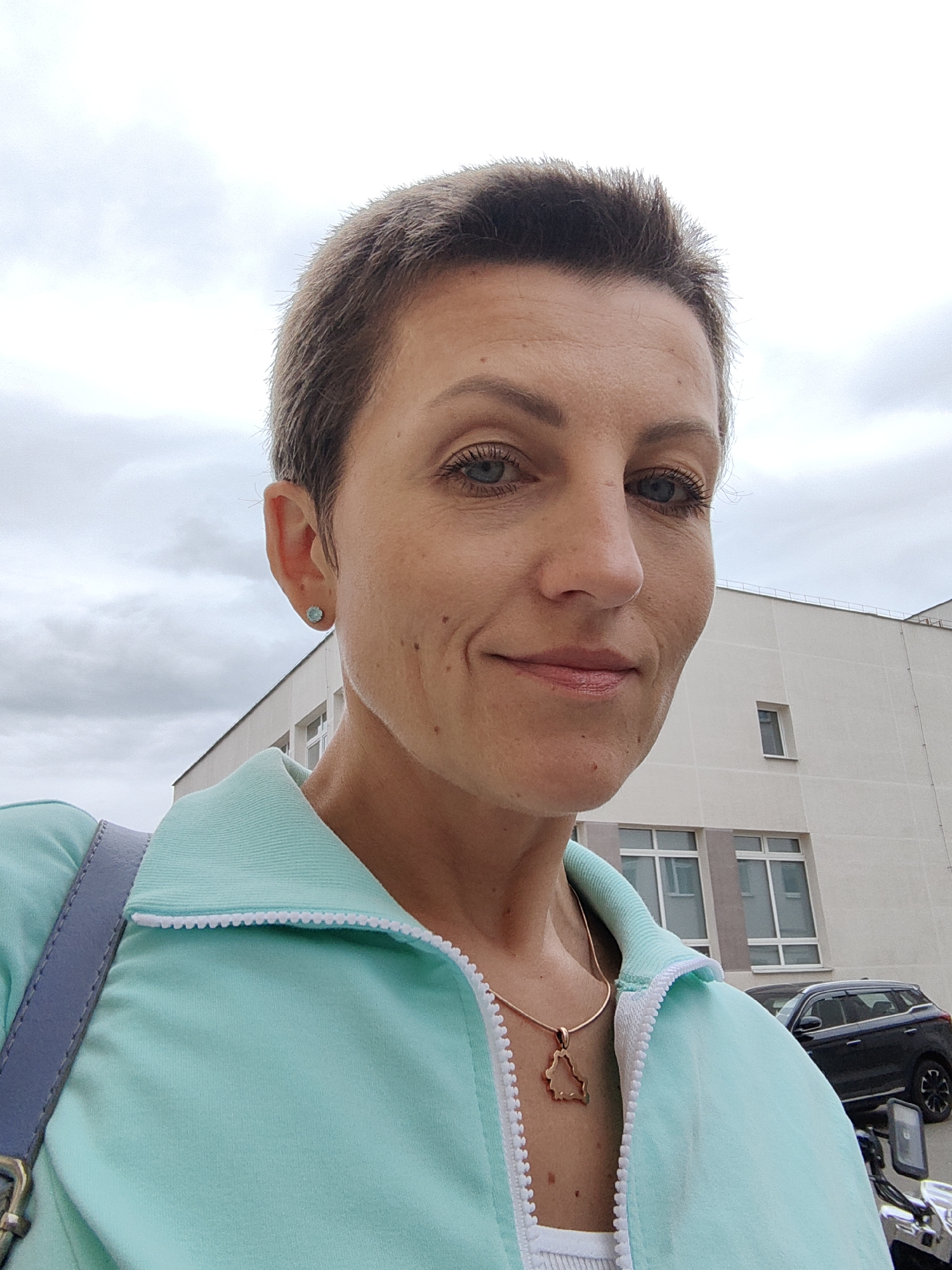 The image is a selfie of a woman with short, brown hair, blue eyes, and light skin. She is wearing a light turquoise jacket with white trim and a white tank top underneath. She has on a delicate gold necklace with a pendant shaped like the outline of Belarus. She is also wearing small blue stud earrings. She is standing outside with a building in the background, which appears to be a modern, light-colored office or residential building with multiple windows. A dark-colored SUV is parked in front of the building. The sky is overcast and cloudy. The overall impression is a casual outdoor portrait. The user mentioned the location is the city of Minsk, Belarus, which is consistent with the country outline pendant.