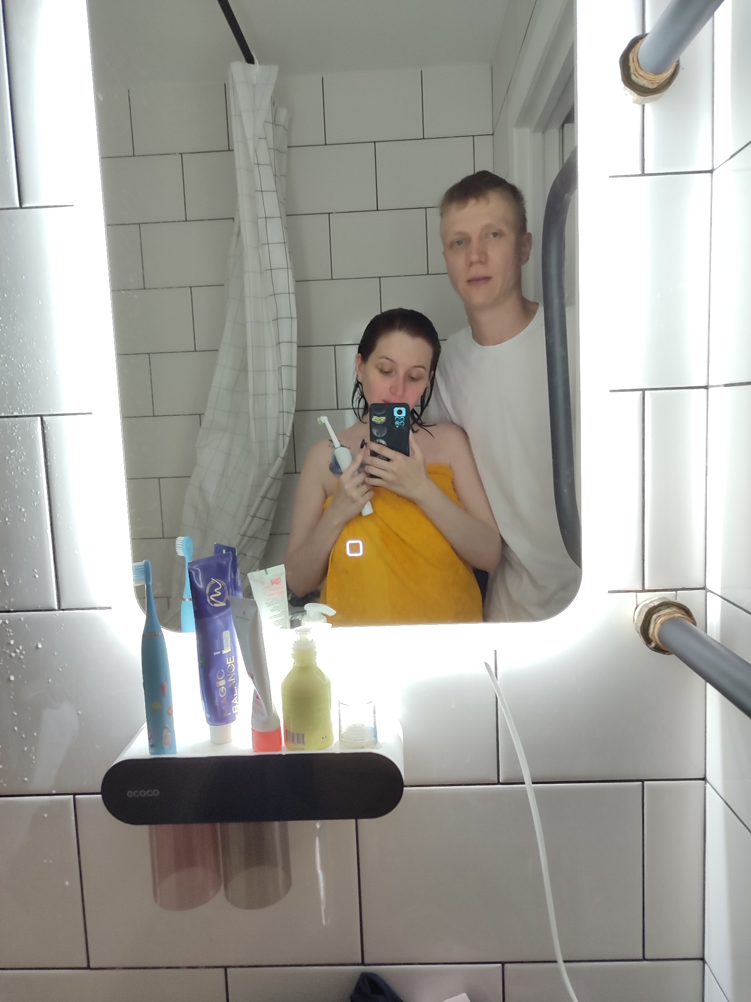 In a bathroom with white subway tile walls, a man and a woman are reflected in a large, well-lit mirror. The woman, in the foreground, is holding a smartphone to take a selfie and is wrapped in a yellow towel. She is holding an electric toothbrush in her right hand and appears to be brushing her teeth. She has dark hair and is looking down at her phone. Beside her, a man with short blonde hair and wearing a white t-shirt stands slightly behind her, looking towards the camera. On the shelf below the mirror, toothbrushes and toiletries are visible, including a blue electric toothbrush, a tube labeled "MAGIC BALANCE," and a yellow dispenser. A white shower curtain with a grid pattern hangs in the background. The overall scene suggests a domestic setting, likely a home bathroom, in Chaikovsky, Russia.