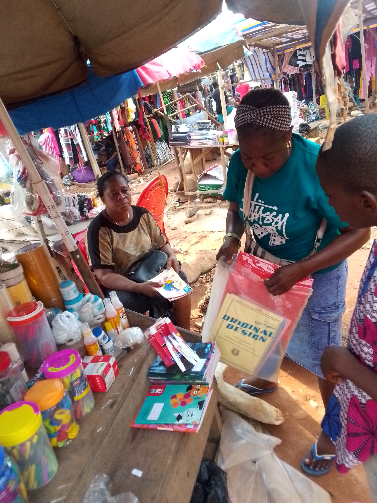 The image captures a scene in an outdoor market, plausibly in Orlu, Nigeria, judging by the style of clothing and the general environment. A woman wearing a green Stussy t-shirt and blue denim skirt is engaged in a transaction. She is reaching into a red plastic bag, revealing a book titled "ORIGINAL DESIGN" inside. Another person, likely a customer, partially appears in the right of the frame, wearing a colorful pink and blue patterned garment. A woman sits on a red plastic chair near a table of goods. On the wooden table are containers filled with colorful items. The background shows a crowded market environment with stalls displaying clothing. The environment indicates a bustling commercial space, with makeshift coverings providing shade.