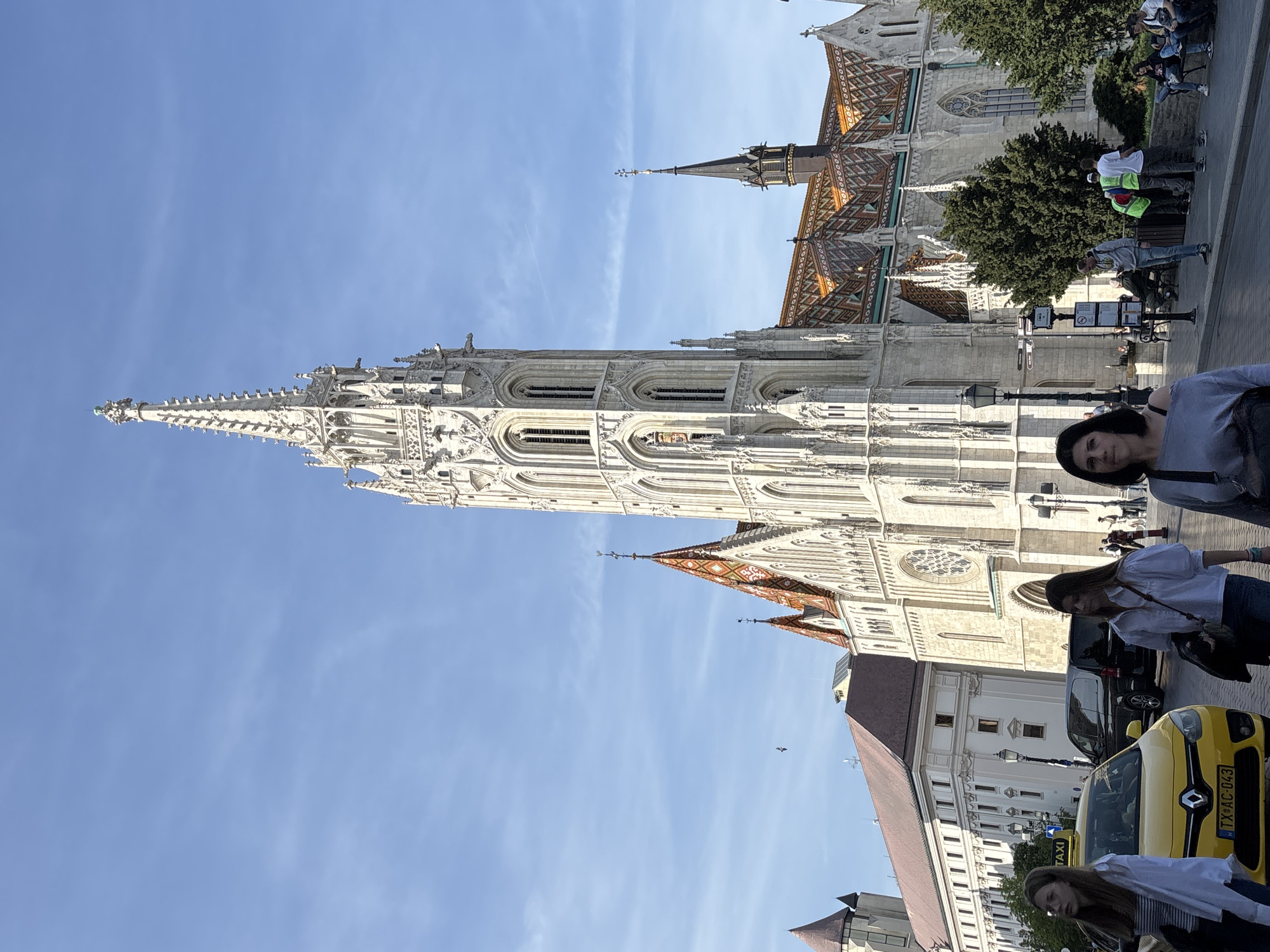 The image displays a bustling outdoor scene dominated by a grand, ornate Gothic-style church under a clear blue sky. The most prominent feature is the church's tall, slender spire, intricately detailed with numerous decorative elements and finials, crafted from light-colored stone. The church also features sections of a distinctive, multi-colored tiled roof, exhibiting geometric patterns.
In the foreground, a woman with dark hair and a dark top is visible on the right, looking towards the camera. To her right, a yellow Renault taxi is prominent, displaying the Hungarian license plate "TX-AC-043" with the country code "H" and the word "TAXI" on its roof light. A dark SUV is parked behind the taxi. Further into the scene, another woman in a white shirt and dark pants is walking. In the middle ground, several people are scattered, including two individuals wearing white shirts and bright green/yellow high-visibility vests near a bench, and others walking or standing. Trees are visible providing shade and greenery near the base of the church.
The setting appears to be a public plaza or street alongside the historic building, likely a popular tourist spot. The time of day is bright and sunny, indicated by the clear blue sky and well-defined shadows, suggesting late morning or early afternoon. The architecture, particularly the patterned roof tiles and the intricate stonework, strongly suggests the Matthias Church in Budapest, Hungary. No specific event is taking place; people seem to be going about their daily activities or sightseeing.