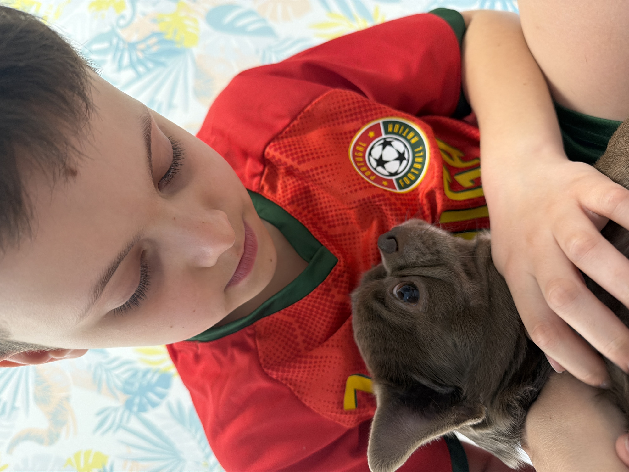 A young boy and a puppy share a tender moment indoors, set against a brightly patterned background, in Votkinsk, Russia. The boy, who appears to be a child or young teenager with short brown hair, is lying down or reclining, gently looking downwards or resting with his eyes closed. He is wearing a vibrant red and green sports jersey. On the chest of the jersey, a circular emblem is visible, featuring the words "PORTUGAL," "FOOTBALL," and "NATION" surrounding a football graphic. He is cradling a small, greyish-brown puppy, which gazes up towards the boy with dark, expressive eyes. The background is a sheet or blanket adorned with a cheerful pattern of light blue, yellow, and white tropical leaf designs. The image conveys a sense of warmth, affection, and peaceful companionship between the boy and his pet. The lighting suggests a bright, daytime indoor setting.