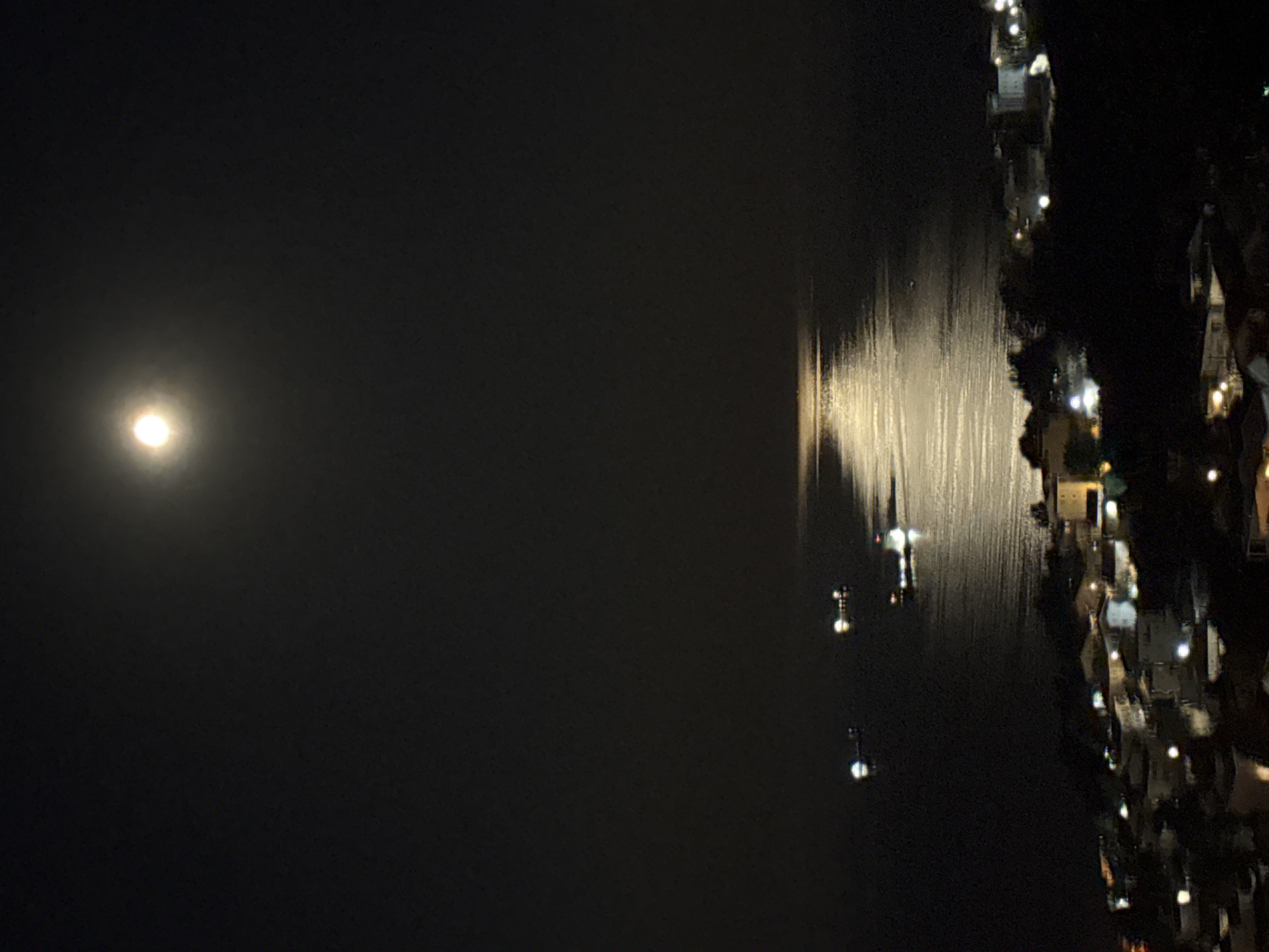 The image shows a nighttime scene in Šušanj, Montenegro. The moon is visible in the upper left. Its reflection is seen in the water in the center of the image. Along the right edge of the image, there are buildings and trees. Lights from the buildings are visible and reflect in the water.