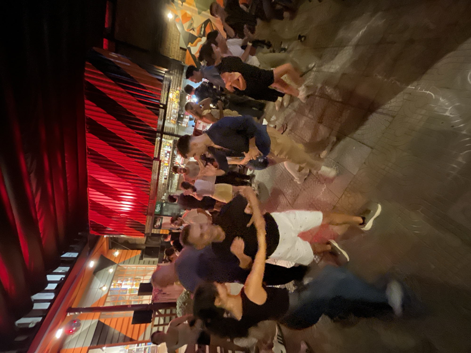 The image depicts an active outdoor or semi-outdoor social venue at night in Đà Nẵng, Vietnam, populated by approximately 15-20 adults. In the central foreground, a man wearing a black short-sleeved shirt and white patterned shorts, with dark sneakers, is engaged in a partner dance with a woman who has dark hair tied back, dressed in a dark sleeveless top and dark pants, also wearing sneakers. Their arms are extended, holding each other's hands. Surrounding them, other individuals are observed dancing, standing, and socializing. The environment features a distinct overhead installation of diagonally striped red and black corrugated material. To the left, a building wall is adorned with angular orange, black, and white geometric patterns, with windows revealing interior shelving and a small disco ball positioned high. To the right, a bar area is visible, stocked with numerous bottles behind a counter, and a Carlsberg logo is faintly discernible on equipment behind the bar. The ground is paved with various tiled patterns, and the scene is illuminated by a mix of red ambient lighting and localized white/yellow lights, casting multiple shadows.