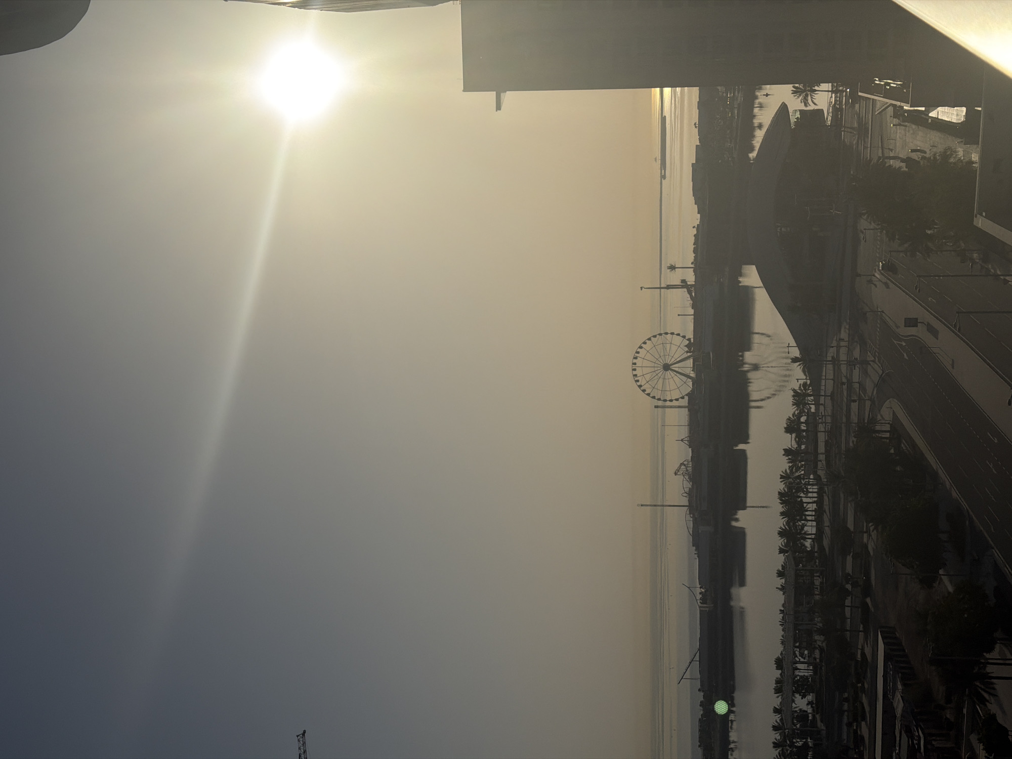 The image depicts a hazy, sunlit cityscape with a body of water in the mid-ground and a distant horizon. A large Ferris wheel is visible on the waterfront, along with other amusement park structures. Several buildings and palm trees are present in the foreground and mid-ground. The sun is bright and positioned in the upper right quadrant of the image, casting a lens flare across the sky. A portion of a building is visible on the right edge of the frame. The overall impression is a coastal urban environment during daylight hours, possibly early morning or late afternoon due to the low angle of the sun and the haziness.