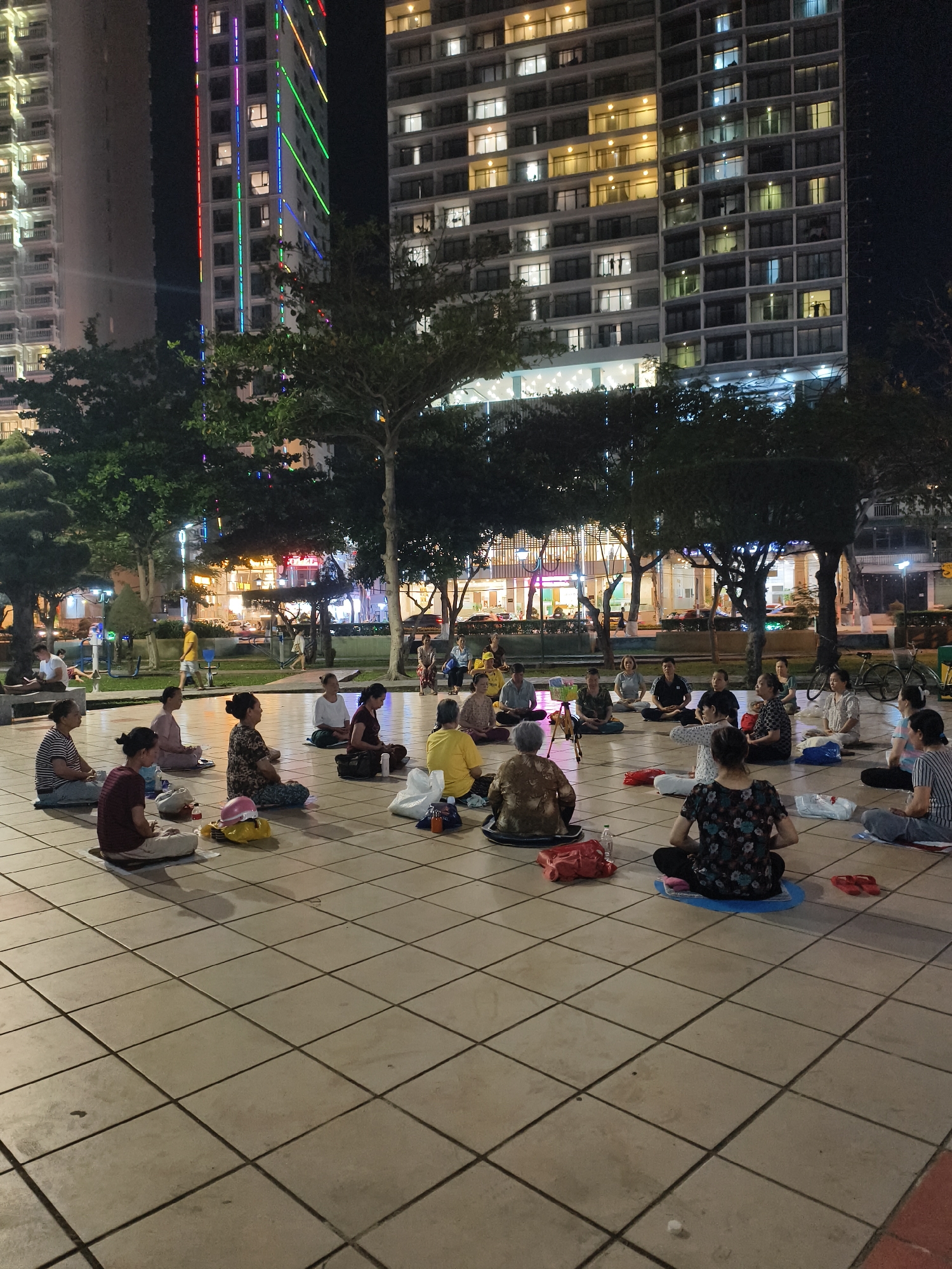A gathering of approximately 20-25 people, mostly women, are seated in a circle on a large, tiled plaza at night. They appear to be meditating or engaging in a group activity that involves sitting cross-legged. Some individuals have mats or cushions beneath them, and there are various personal items like bags and water bottles scattered around.
The location is a public space within a city; tall buildings with illuminated windows serve as a backdrop, including one with colored lights running vertically along its facade. Trees surround the plaza, providing some shade and contributing to the urban park setting. In the background, there's a hint of street activity, with lights and movement suggesting the presence of businesses or public amenities nearby. The setting is Unknown, Unknown.