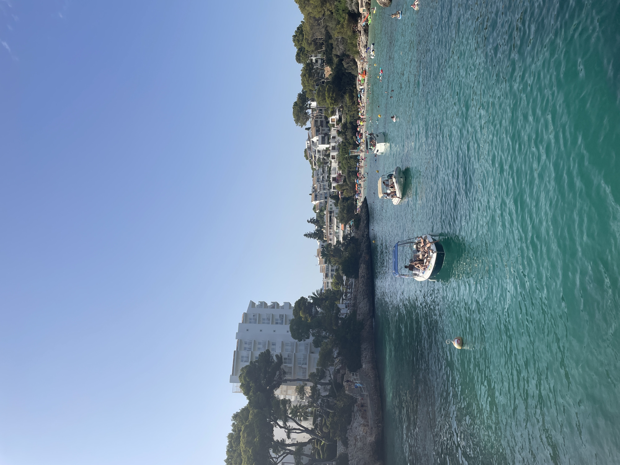 A wide-angle, eye-level shot captures a serene bay at Cala d'Or, Felanitx, Spain, bathed in the clear light of day. The foreground is dominated by the tranquil, turquoise waters of the Mediterranean Sea, with gentle ripples disturbing the surface. A solitary, orange and white buoy bobs near the center of the frame.
Further into the bay, two small boats navigate the water. The boat closest to the viewer, on the right, has its front facing left, carrying at least three individuals sunbathing on its bow. The second boat, situated to the left and slightly further back, is also carrying a group of people, some appearing to be seated.
Beyond the boats, the scene opens up to a vibrant beach bustling with activity. Numerous beachgoers are scattered across the sandy expanse, some under colorful umbrellas, others wading in the shallow water. The shoreline is lined with lush green trees and rocky outcrops.
Dominating the left side of the background is a large, multi-story white hotel complex, its balconies adorned with railings. To the right of the hotel, a cluster of white residential buildings cascades down the hillside, interspersed with trees. The sky above is a vast expanse of clear, pale blue, devoid of clouds. The overall impression is one of a popular tourist destination, alive with the activities of a sunny summer day.