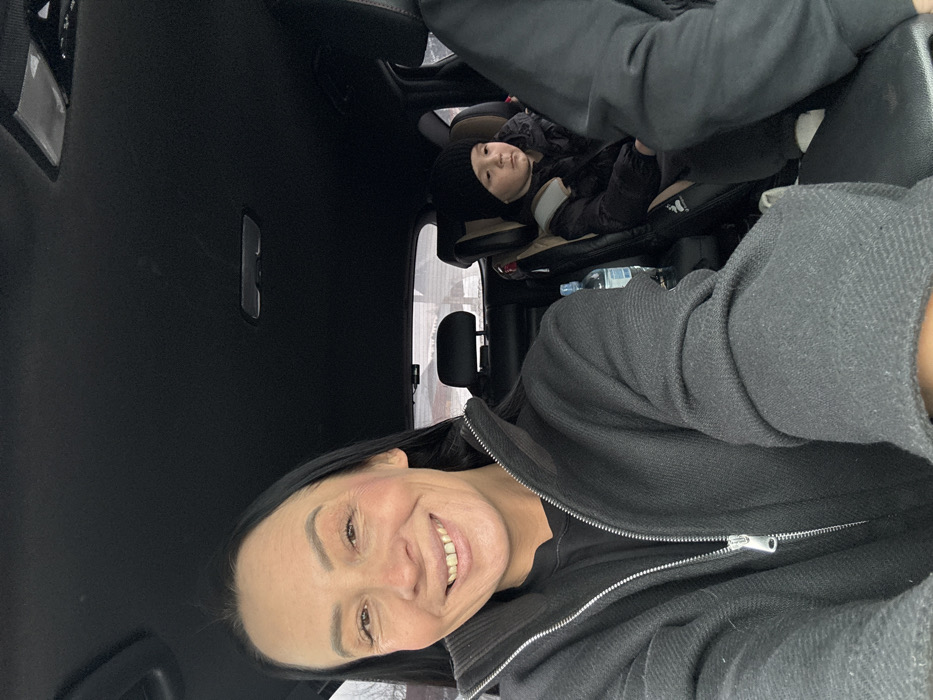 The image depicts the interior of a vehicle, presumed to be in Pavlovsky, Russia, during what appears to be cold weather. A woman occupies the foreground, positioned to the left. She has long, dark hair and is smiling directly at the viewer, revealing her teeth. She wears a dark, zip-up jacket over a dark shirt. To the right, in the midground, a young child is seated in a rear-facing or forward-facing child safety seat. The child wears a dark knitted beanie and a dark puffer jacket, looking with a neutral expression towards the right side of the frame. A plastic water bottle with a blue label is visible next to the child's seat. A partial view of a third individual's dark-sleeved arm and shoulder is visible in the extreme foreground on the right. The vehicle's interior features a dark headliner and dark upholstery. Through the rear window, an overcast sky and a barren or snow-dusted landscape are visible.