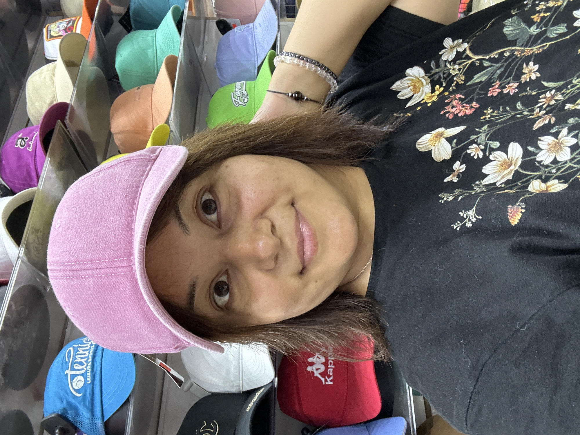 A young woman with brown hair is taking a selfie in front of a display of hats. She is wearing a pink baseball cap and a black t-shirt with a floral print. The hats are displayed on shelves in a store. The woman is smiling and looking at the camera. There are no other people visible in the image. The environment appears to be a retail store.