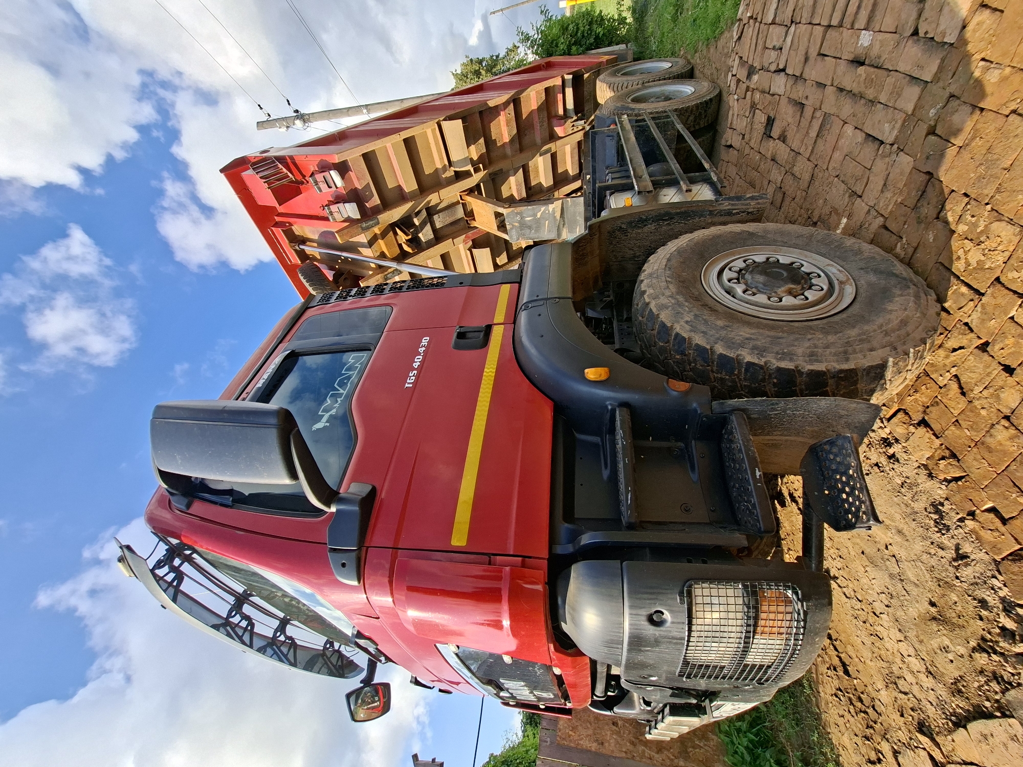 An exterior, low-angle, wide shot captures a bright red dump truck, positioned on a dirt and brick road. The truck's bed is elevated, suggesting it is either loading or unloading. The sky above is a vivid blue, dotted with fluffy white clouds. To the right of the truck, a utility pole with wires is visible, and further to the right, green foliage and trees line the background. The front of the truck is facing left, with its large tires prominent in the foreground, indicating a robust and heavy-duty vehicle. The overall impression is one of a working vehicle in an outdoor, possibly rural or construction, setting.