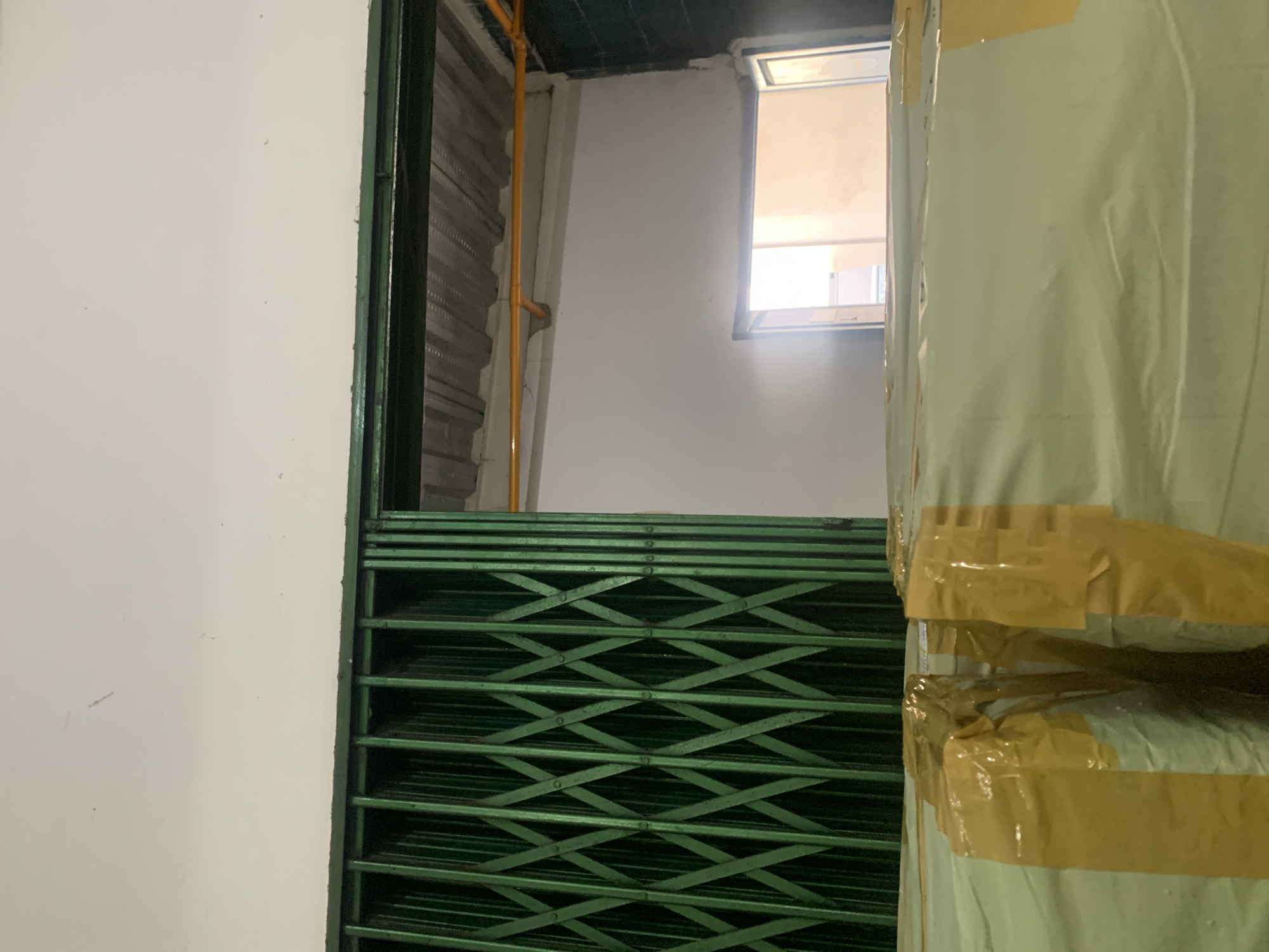 The image shows the interior of what appears to be a storage facility or warehouse in Palembang, Indonesia. In the foreground, several large packages wrapped in light-colored material and secured with yellow tape are visible. A green metal accordion gate dominates the left side of the frame, partially obscuring the view into an adjacent space. Beyond the gate, a corrugated metal roof is visible. To the right, a doorway opens into another area, illuminated by natural light from an unseen source. A yellow pipe runs across the ceiling in the background. The walls are painted white.