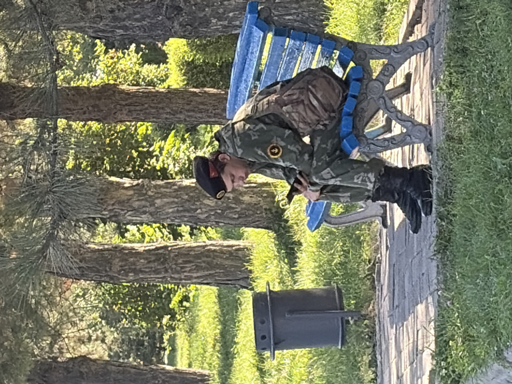 A young man wearing a military uniform, including a camouflage jacket, trousers, black boots, and a black beret, is sitting on a blue park bench. He is looking down at his smartphone. The man has a brown shoulder bag slung across his chest. He is in a park-like setting with trees and grass. To his left, there is a dark-colored trash can. The bench is ornate with a grey metal base and blue wooden slats. The ground in front of the bench is paved with light-colored bricks. The lighting suggests it is daytime, with dappled sunlight filtering through the trees.
