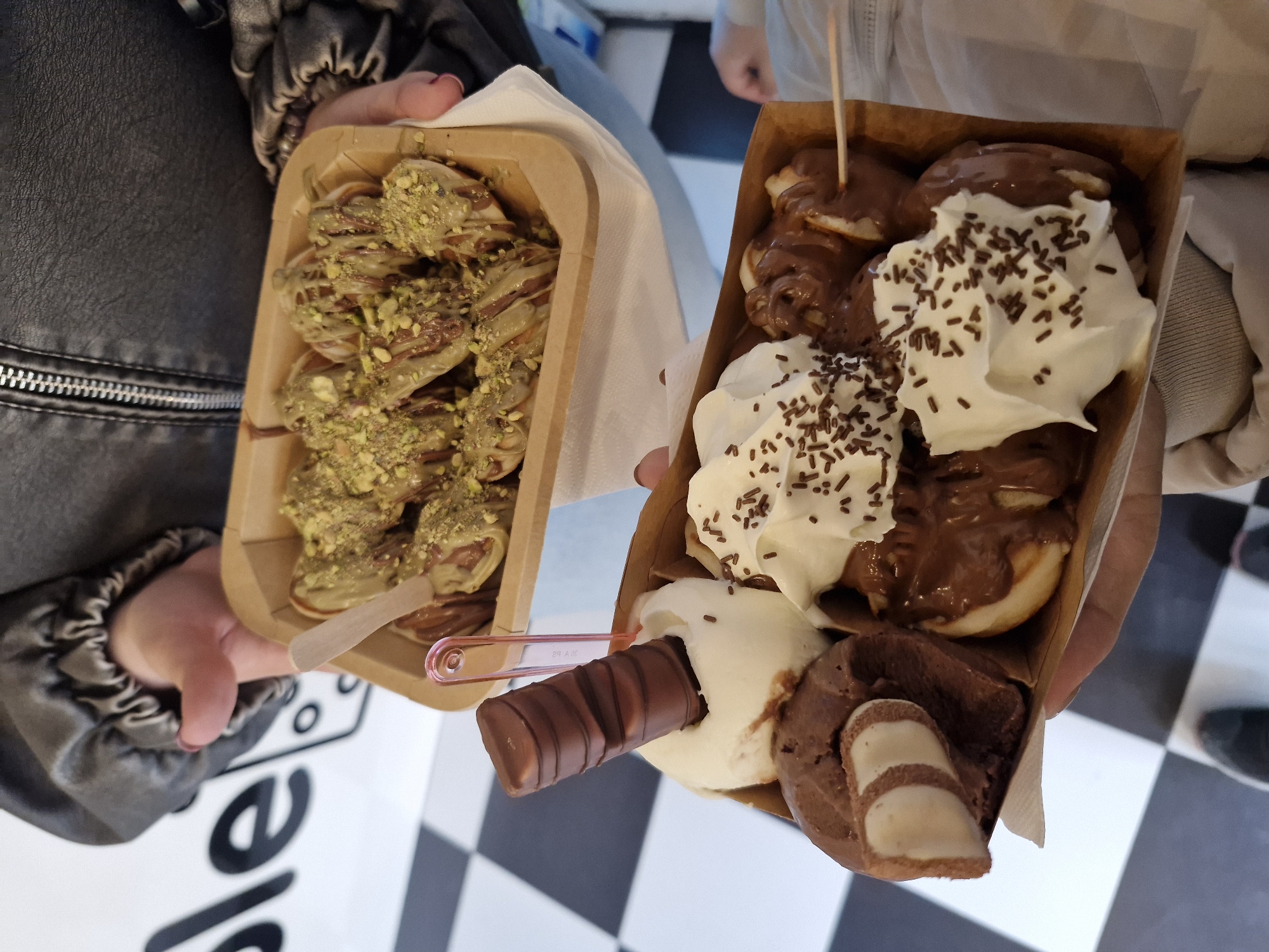 This close-up, high-angle image captures two individuals holding elaborate dessert boxes, likely taken indoors at a dessert shop or cafe in Sofia, Bulgaria.
On the left, a person wearing a black leather jacket with a prominent zipper on the sleeve holds a rectangular cardboard container. Inside are numerous small, rolled-up pastries, possibly mini crepes or pancakes, generously covered in a light green sauce, likely pistachio-flavored, and sprinkled with crushed pistachios. A small, light-colored wooden spoon rests on the side of the container.
On the right, another person, dressed in a light-colored jacket or hoodie, holds a similar cardboard box filled with a different selection of sweets. This box contains several mini pancakes or puffy pastries, smothered in dark chocolate sauce and topped with multiple dollops of whipped cream and chocolate sprinkles. Additionally, there's a scoop of white ice cream, a scoop of chocolate ice cream with a segment of a Kinder chocolate bar, and a whole Kinder Bueno bar. A pink plastic spoon and a wooden stick are also visible within this dessert.
The floor beneath the desserts is a distinctive black and white checkerboard pattern. Partial text, specifically the letters "e" and "le", can be seen on the white tiles of the floor, possibly part of a brand name or decorative signage. The overall scene suggests a moment of indulgence and enjoyment of sweet treats.
