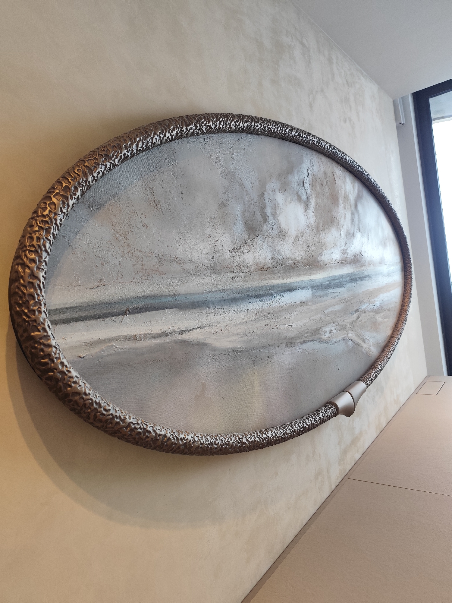 The photograph depicts an oval-shaped piece of art affixed to a wall. The art piece has a textured, bronze-colored frame that encases an abstract painting with a predominantly grey, white, and blue color palette. The painting evokes a landscape scene, possibly a beach or shoreline, with horizontal lines suggesting the division between land, sea, and sky. The wall behind the artwork is textured with a mottled beige finish. To the right of the artwork, there is a smooth, light brown surface, possibly a countertop or the top of a cabinet. A window is visible in the upper right corner, suggesting natural light illuminating the scene. The setting appears to be an indoor space, likely a room in a building in the city of Unknown, Unknown. No people are visible in the image.