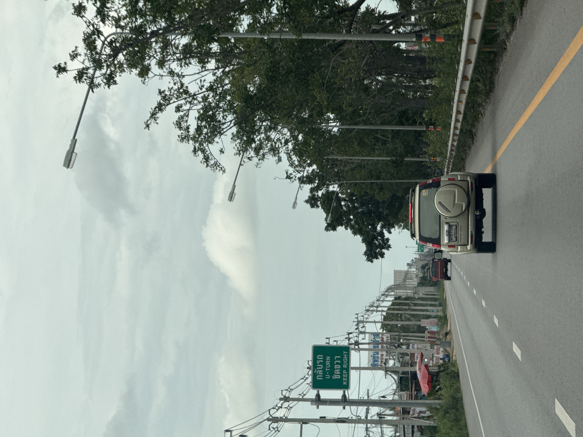 The image captures a view from inside a vehicle traveling on a multi-lane road under an overcast sky. In the foreground, the rear of a silver SUV with license plate "409 7135" is visible. To the left of the road, there are utility poles with numerous wires, a green road sign indicating "U-TURN" and "KEEP RIGHT", and some small buildings and businesses, including a red umbrella. Further down the road, a red SUV is also traveling. The right side of the road features a metal guardrail and a dense line of trees. The overall environment suggests a semi-urban or suburban setting. The green sign indicates this location is in the vicinity of Ban Nong Bua, Thailand.
