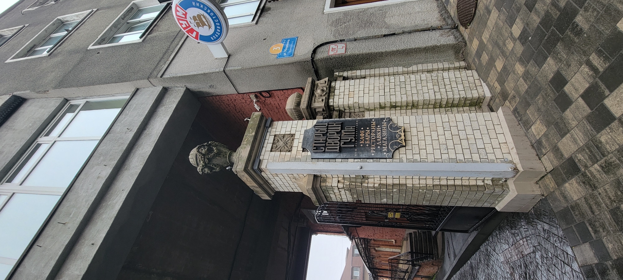 The image shows the entrance to a courtyard area in Vladivostok, Russia. A sign on a brick pillar reads "СТАРЫЙ ДВОРЕЦ ГУМА" which translates to "OLD GUM COURTYARD" and lists services such as "CAFÉ", "BARS", "RESTAURANTS", "SHOPS", and "STORES". The pillar is topped with a decorative urn featuring four faces. To the left of the pillar is a black wrought-iron gate. The building itself is a grey multi-story structure with visible windows. A circular sign with a ship and Russian text is mounted on the building. The ground is paved with interlocking stones. The overall impression is of a historical commercial or mixed-use complex.