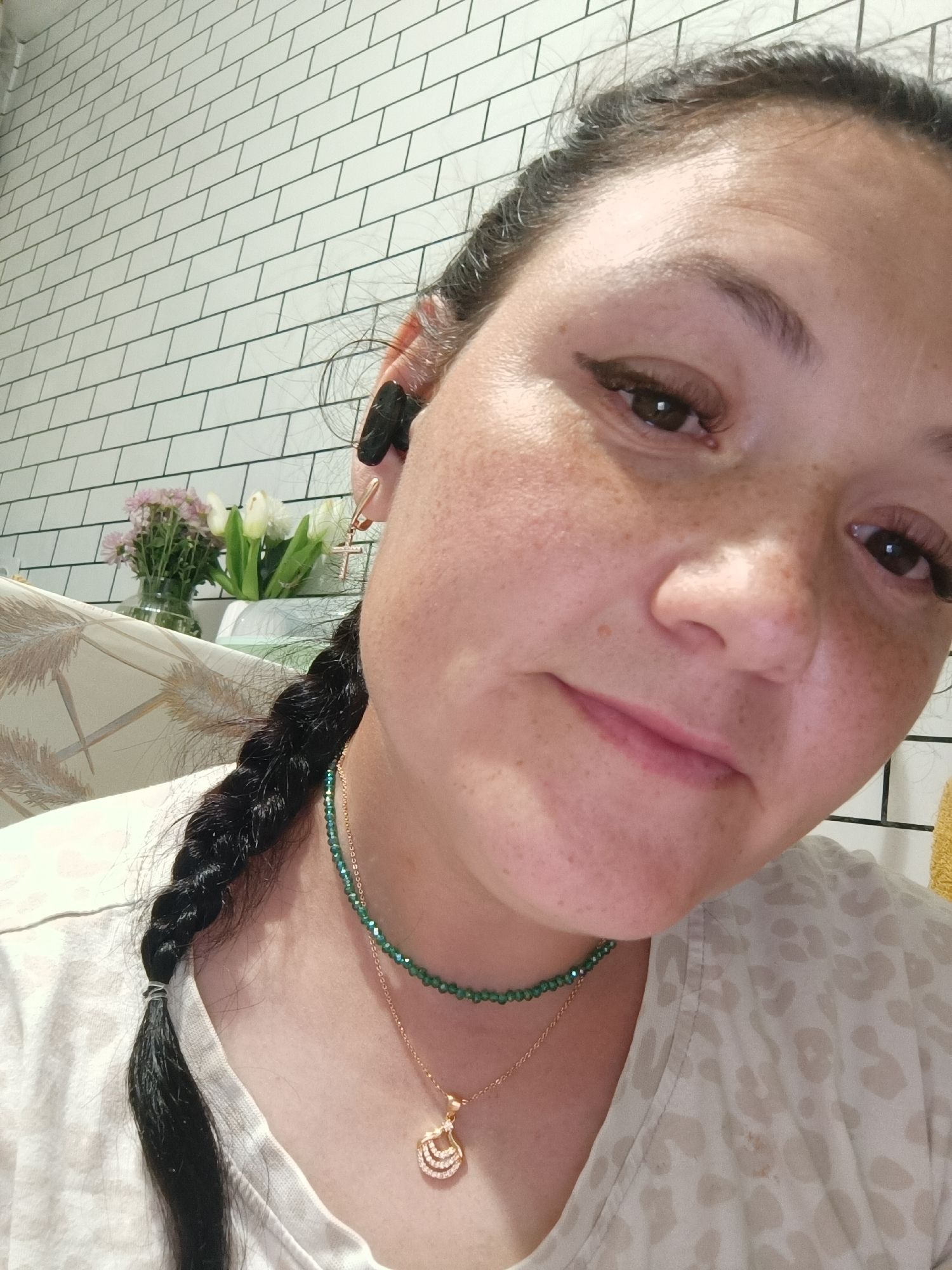 A close-up, head-and-shoulders selfie of a woman is shown, taken from a low angle. She has dark hair braided down her left side, prominent eyelashes, and freckles across her nose and cheeks. She is wearing a light-colored shirt with a subtle leopard print and two necklaces: one a delicate green beaded chain and another with a gold pendant. She has a black earbud in her right ear and a gold cross earring in her left. In the background, a wall of white subway tiles is visible. To the left, out of focus, are a vase of pink and white flowers. The scene appears to be an indoor setting, possibly a bathroom or kitchen, given the tile work. The image does not contain enough information to determine the location within the city of Chaikovsky, Russia.