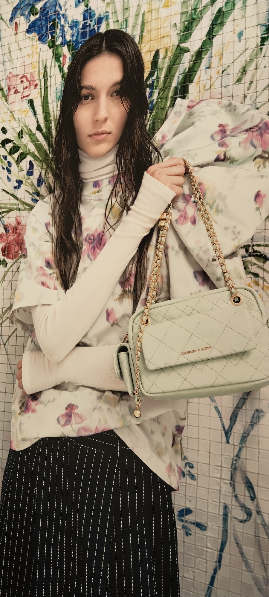 A young woman with long, dark, wet hair stands in front of a colorful, floral-patterned backdrop. She is wearing a white turtleneck, a white top with a floral print, and black pinstripe pants. She is holding a light green quilted handbag with a gold chain strap. The handbag has a gold logo that reads "CHARLES & KEITH". The background appears to be a tiled wall with a painted floral design. There is no indication of the city of Semarang, Indonesia, or any other specific location. The image depicts a fashion-style portrait.