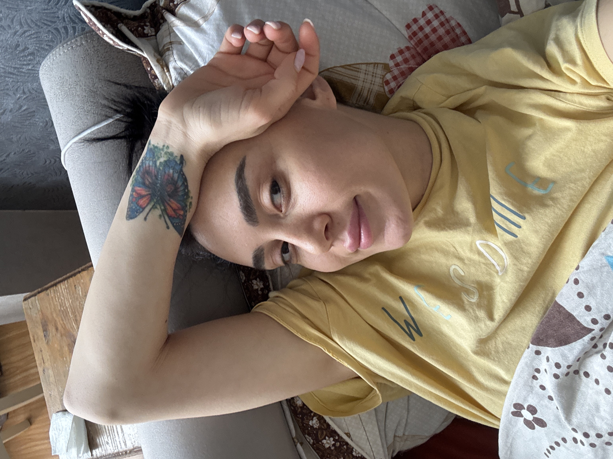 A woman is depicted lying down indoors, viewed from an overhead perspective, with her head oriented towards the bottom right of the frame. She has dark hair and dark, well-defined eyebrows, and is looking towards the camera with a relaxed expression and a faint smile. Her left arm is bent, with her hand resting on her forehead; her fingernails have a light pink polish. A colorful tattoo of a butterfly, featuring shades of red, blue, and green, is prominently displayed on her inner left forearm/wrist.
She is wearing a light yellow, short-sleeved t-shirt with partial text visible on the front. Readable letters include a light blue "W," a light blue "E," and white letters "SD," possibly forming part of a word like "WEDNESDAY." She is resting on a surface that appears to be a couch or bed, covered with patterned fabrics: one section is white with brown floral and dot patterns, and another features a red and white checkered design. A wooden surface, possibly a side table or furniture armrest, is partially visible to her left. The scene suggests a casual, indoor setting, likely during daytime. The provided context indicates the location is Tbilisi, Georgia.