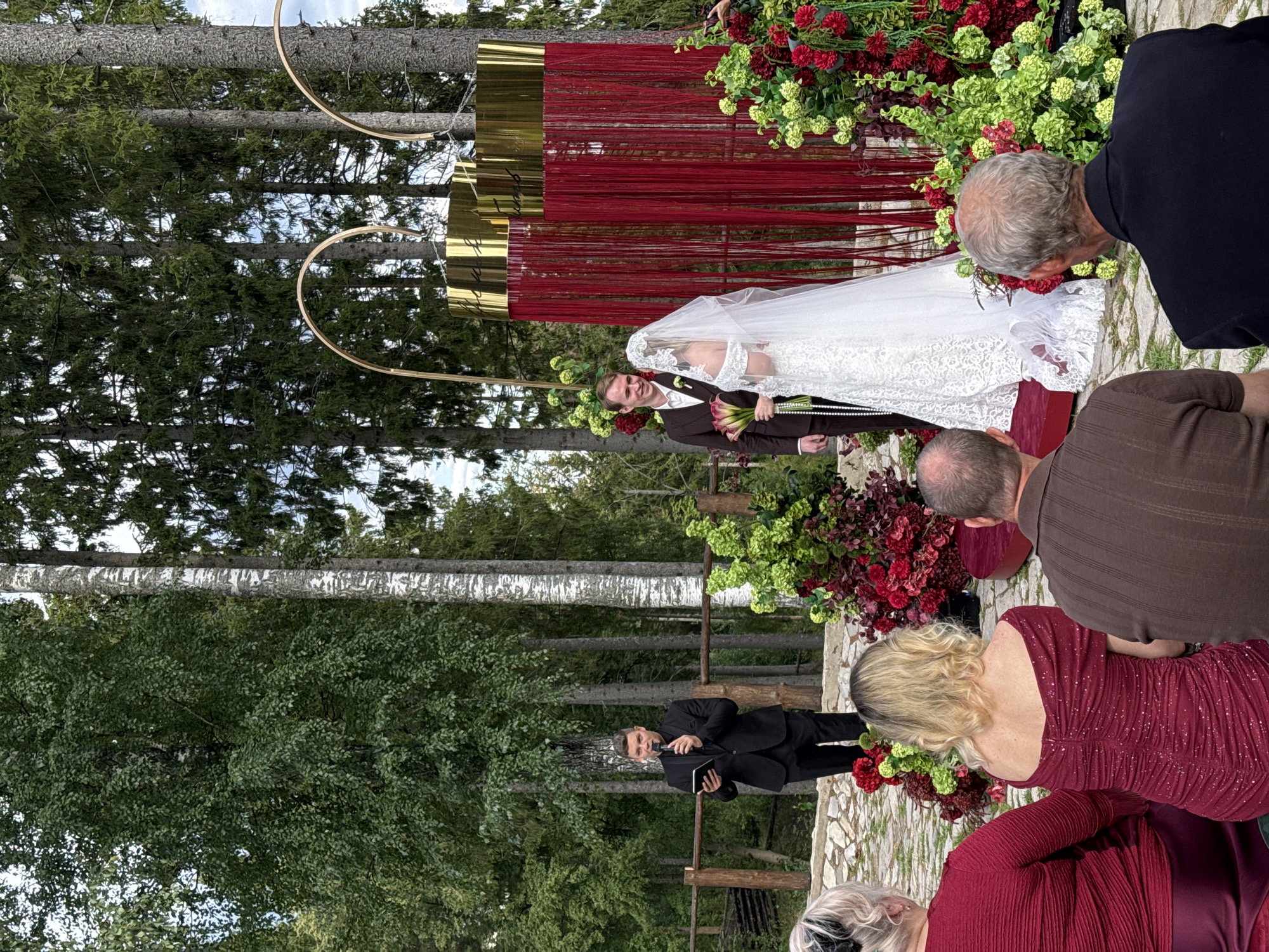 In Kanabekovo, Russia, a wedding ceremony is taking place outdoors amidst a forest setting. A man in a black suit, holding a tablet and a microphone, officiates the ceremony. He stands to the left of a couple, a man in a brown suit and a woman in a white wedding dress, positioned in front of a decorative archway adorned with red fringe and gold accents. The couple is surrounded by elaborate floral arrangements featuring red and green flowers. Several guests are seated in the foreground, facing the ceremony, with two women in red dresses and two men in suits. The environment is characterized by tall trees and a natural, rustic landscape.