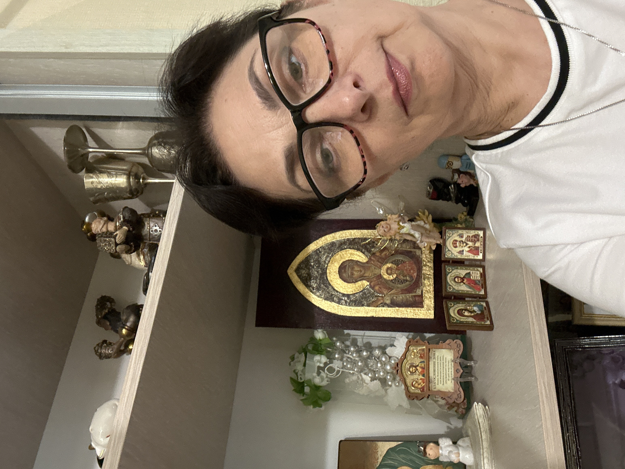 The image depicts a woman wearing glasses and a white t-shirt, looking towards the camera with a neutral expression. She is positioned in the foreground, with shelves filled with various religious icons and small figurines visible behind her. The shelves appear to be made of light-colored wood. Among the objects on the shelves are several icons depicting religious figures, a golden chalice, and a white decorative floral arrangement with pearls. A small angel figurine is also visible. The background consists of a plain wall. The lighting suggests an indoor setting. The image appears to have been taken in a home or a place of worship. There is no specific indication of the city of Hnarovske, Ukraine.