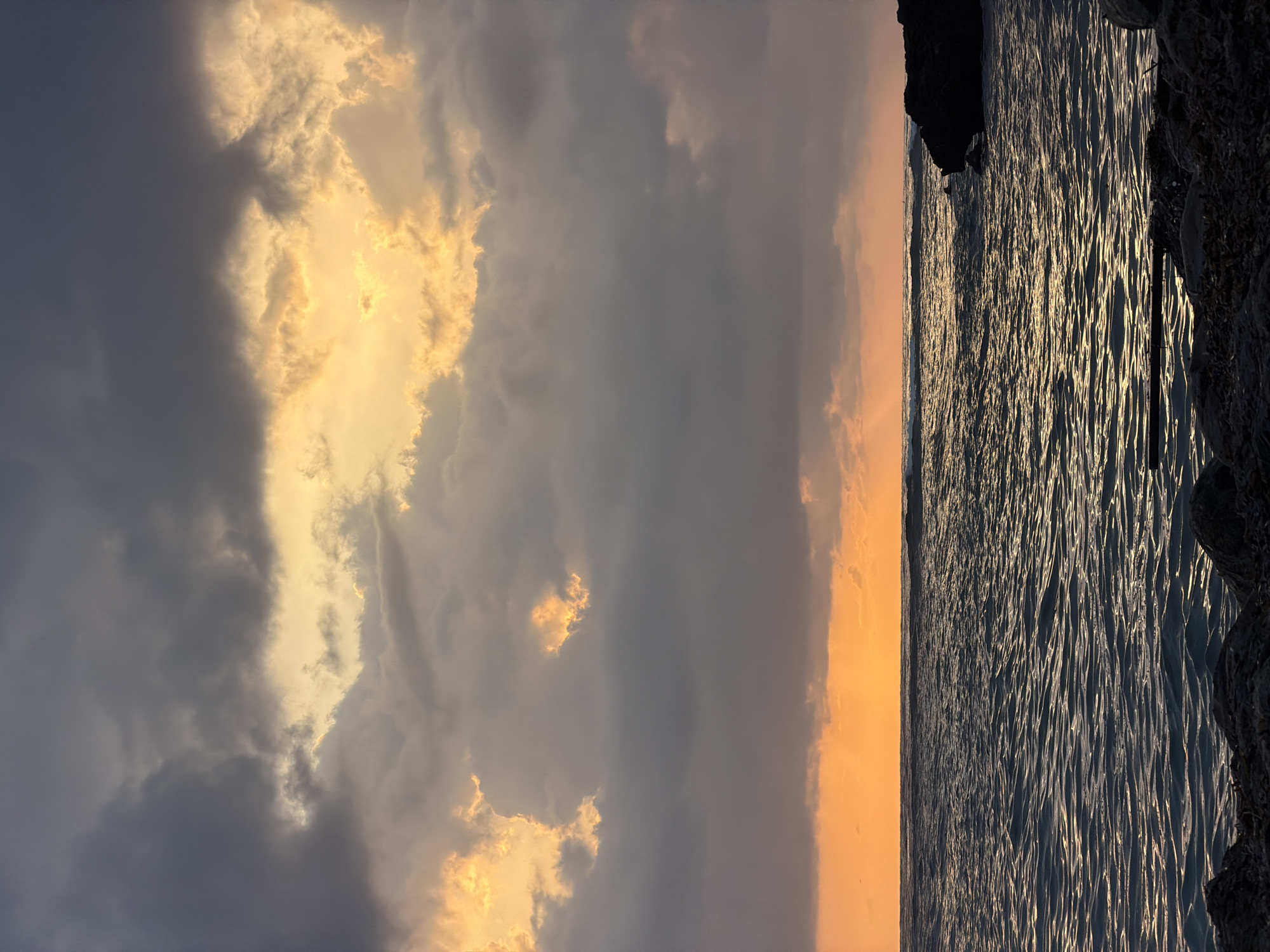 The image depicts a coastal scene with a dark, choppy ocean under a dramatic sky filled with clouds. The sky is a mix of dark grays and vibrant oranges and yellows, suggesting a sunset or sunrise. Light rays are visible breaking through the clouds over the ocean. In the foreground, there are rocks and some debris along the shoreline. The water reflects the colors of the sky, creating shimmering patterns. There are no people or specific structures visible. The location is described as Rotes, Spain.