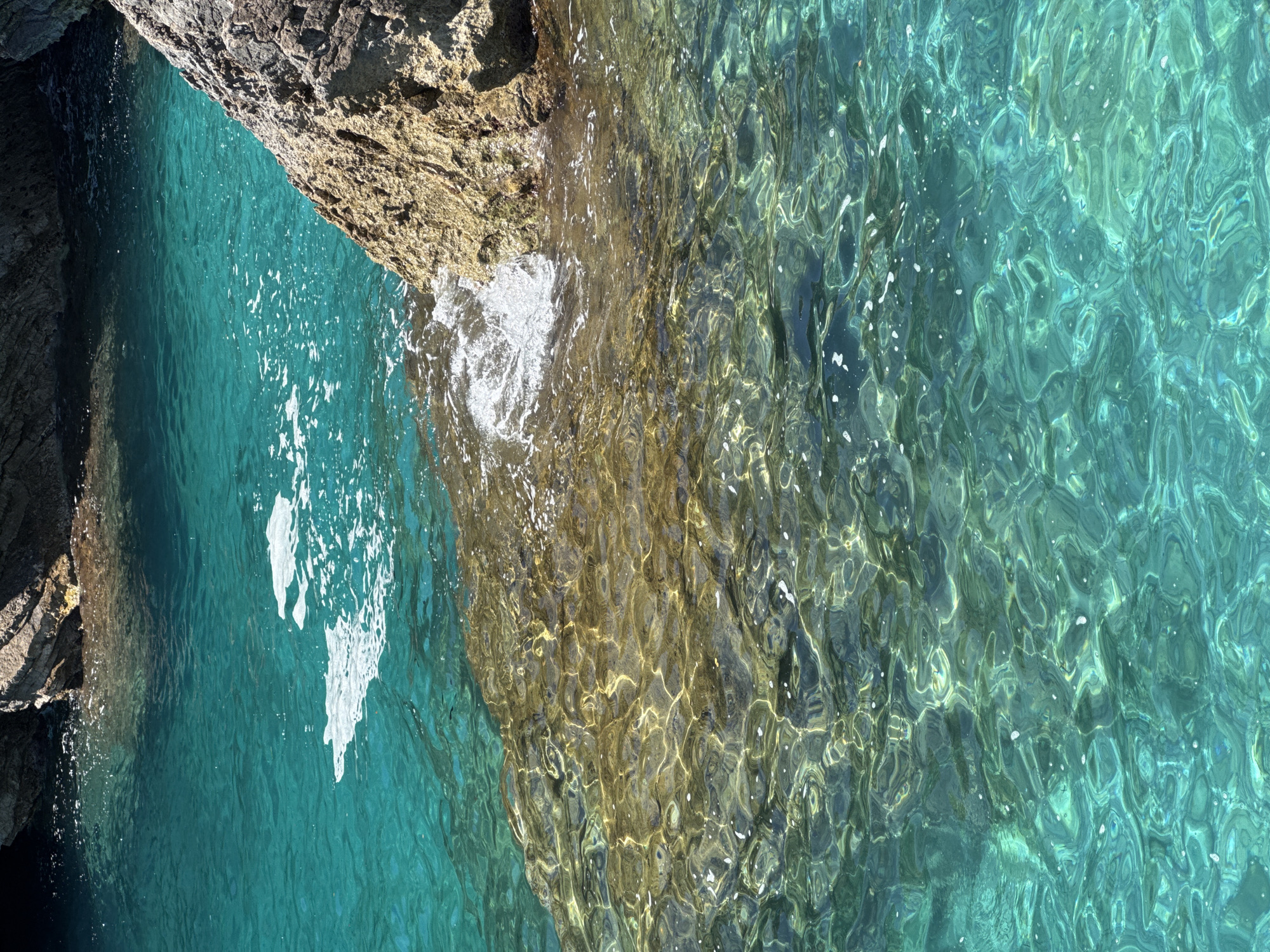 This high-angle shot captures a pristine coastal scene in Budva, Montenegro, featuring a striking contrast between rugged rock formations and luminous, clear water. The image is dominated by the sea, displaying an exquisite gradient of turquoise, teal, and emerald green, reflecting varying depths and light conditions.
On the left side, a dark, textured rock cliff descends sharply into the water, creating a shaded area of deeper blue. Along the upper edge and extending into the middle, lighter, golden-brown rock formations are visible, some partially submerged beneath the transparent water. White foam and spray are present where the water gently crashes against these rocks, particularly noticeable on the left and in the center.
The surface of the water is alive with intricate patterns of sunlight, creating dazzling refractions and ripples that reveal the rocky seabed beneath. This clarity indicates very clean water. The bright illumination suggests a sunny daytime, likely mid-day, enhancing the vibrant colors of the water and rocks. The scene evokes a sense of natural beauty, tranquility, and the pristine quality of a coastal environment. There is no visible text in the image.