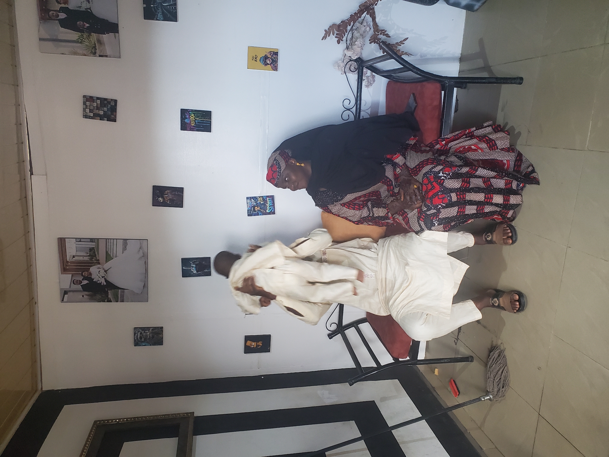 A woman in a dark head covering and patterned fabric sits on a small metal-framed chair. She appears to be interacting with a person in a cream-colored outfit, who is holding a baby. The scene is indoors, with a wall behind them adorned with multiple framed photos and prints. The flooring is light-colored tile. A mop is visible on the floor next to the chair. The location is likely in Jos, Nigeria.
