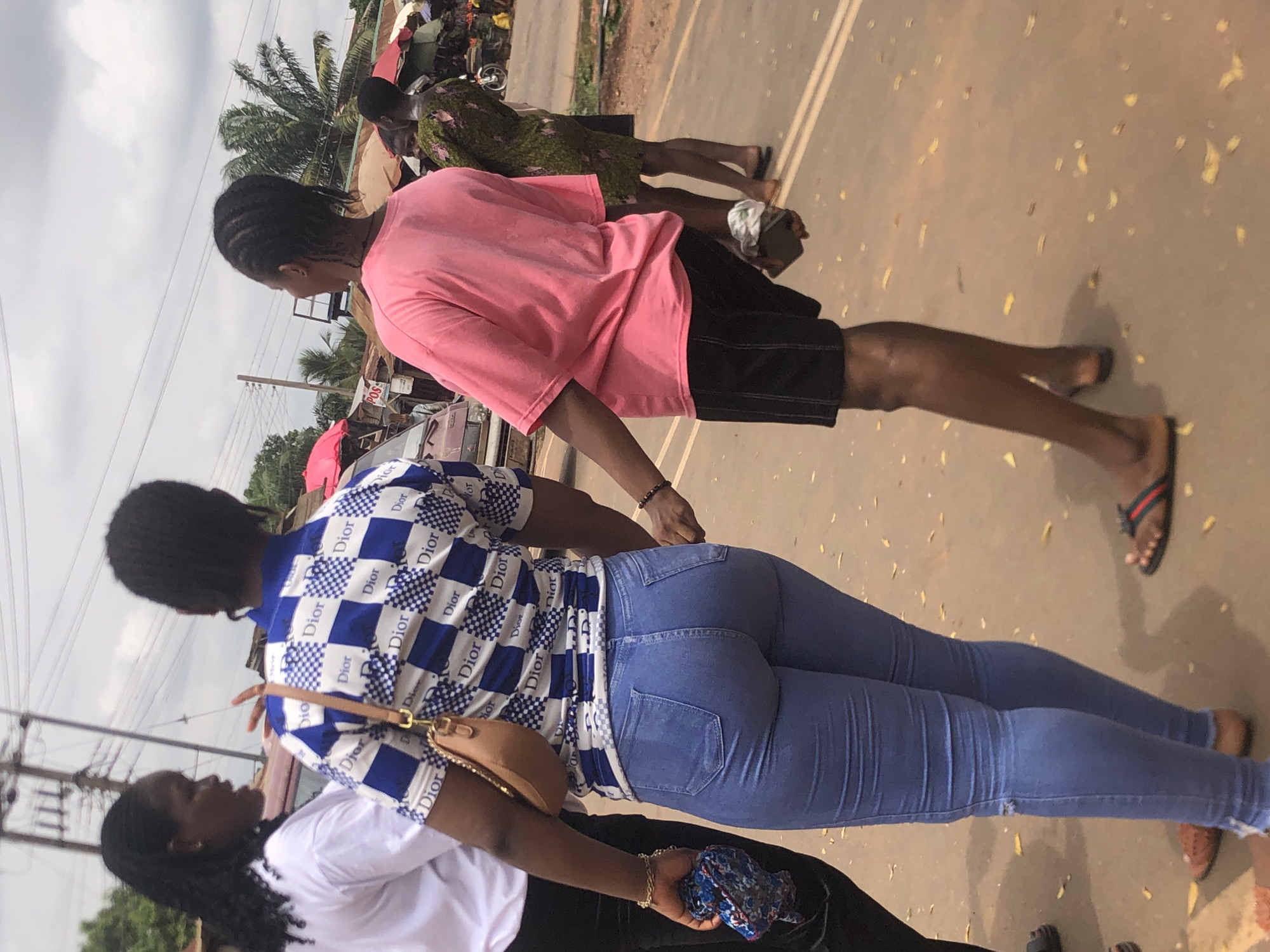 The image captures an outdoor street scene in what appears to be Nsugbe, Nigeria, during daytime under a cloudy or hazy sky. The ground is a light brown, possibly unpaved road or dirt path, with scattered yellowish debris. Overhead, numerous power lines are visible, supported by utility poles, and palm trees line the background.
In the foreground, a young woman is seen from behind, facing away from the camera. She wears a striking blue and white checkered shirt with "Dior" logos repeatedly printed across it, and fitted blue skinny jeans. A tan shoulder bag hangs from her shoulder, and she holds a blue patterned cloth in her left hand. To her left, partially obscured, is another person wearing a white t-shirt and dark pants.
Further back, slightly to the right, another person walks in the same direction, also seen from behind. This individual wears a pink t-shirt, black shorts, and red and black striped flip-flops. They have black braided hair and appear to be holding a dark smartphone in their left hand. In the background, behind this person, another figure in a green patterned dress is visible.
The scene suggests a busy public area, possibly a marketplace or a main thoroughfare. A small white sign with "POS" (Point of Sale) is visible in the background, indicating a commercial establishment. The people appear to be going about their daily activities, walking along the path.