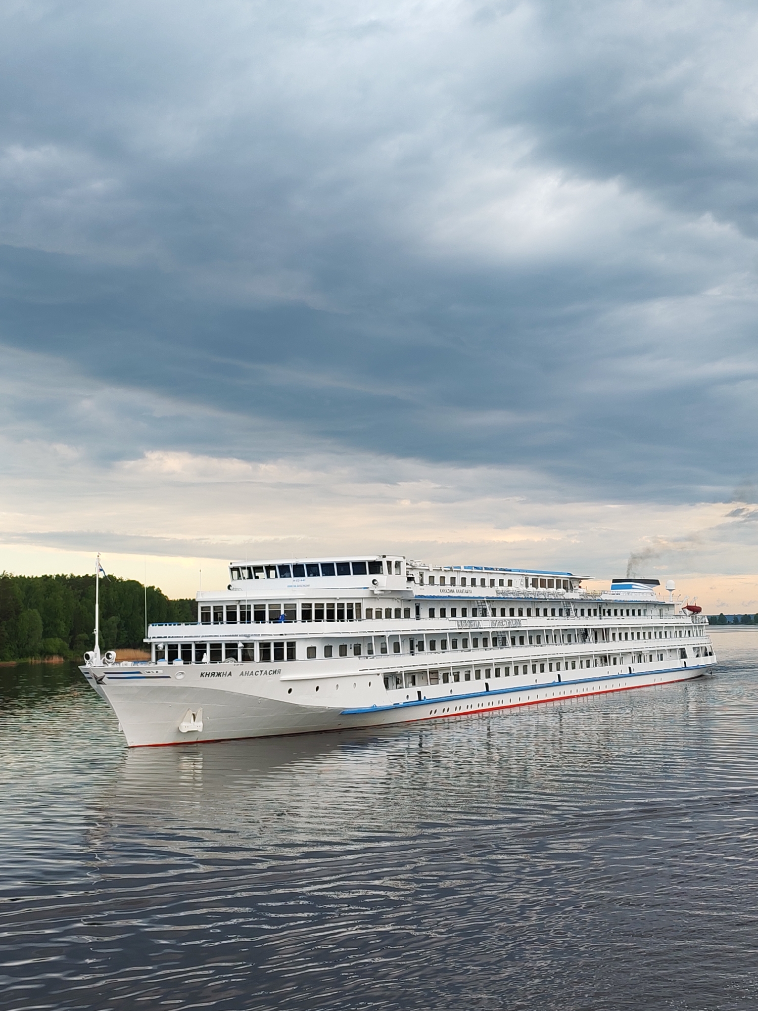 A long, white passenger ship, "KNIAZHNA ANASTASIA," is sailing on a body of water. The ship features multiple decks, numerous windows, and a blue stripe along its lower section. The front of the ship displays the name in Cyrillic script.
The water has a rippled surface, reflecting the sky. On the left, a strip of green forest is visible along the shoreline. The sky is dominated by heavy, dark clouds.
The location is identified as Eremeitsevo, Russia.